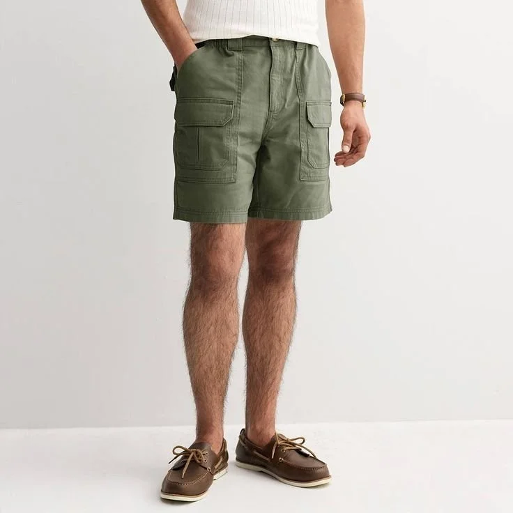 Person wearing green cargo shorts, brown boat shoes, and a white shirt with their left hand in pocket against a plain white wall.