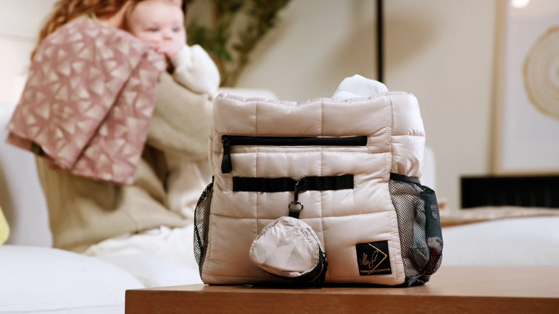 A pink quilted backpack with black zippers and mesh side pockets placed on a wooden table, with a woman holding a child blurred in the background.