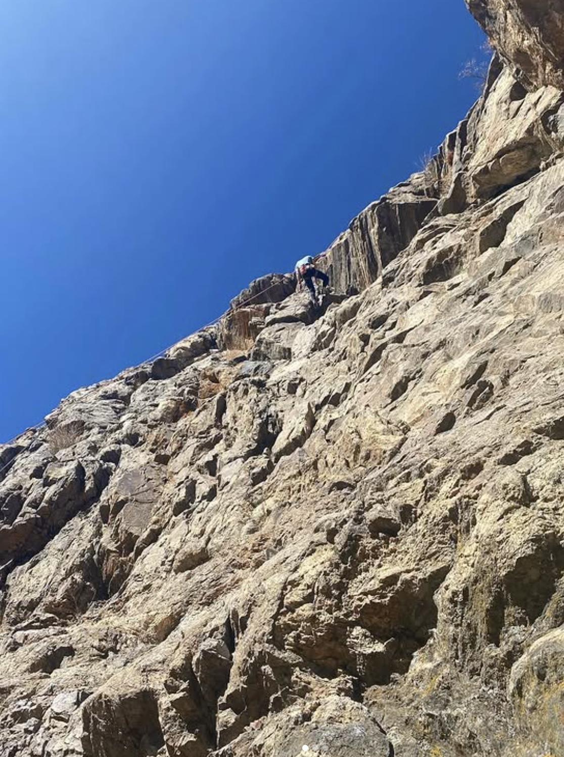 A person climbing a steep, rocky mountain face under a clear blue sky.