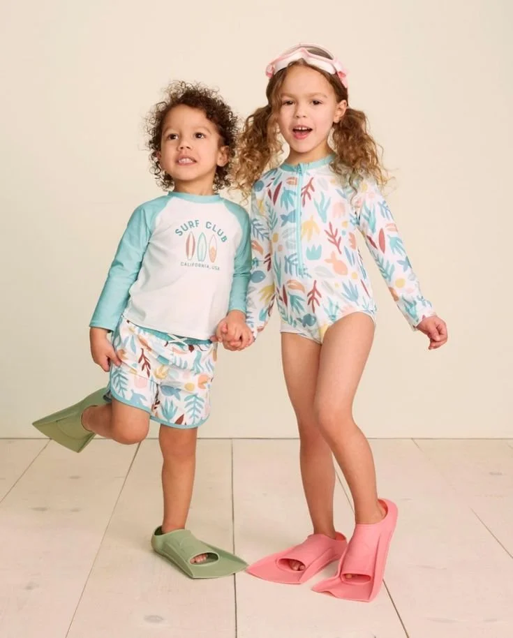 Two young girls holding hands, wearing colorful beachwear and oversized foam slippers, standing on a light-colored floor against a plain wall.
