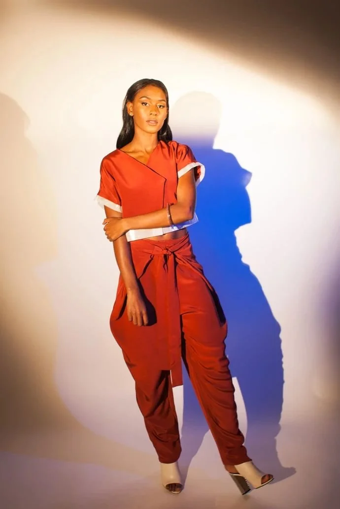 A woman wearing a red outfit composed of a short-sleeve top and matching pants, standing against a plain background with dramatic lighting creating shadows.