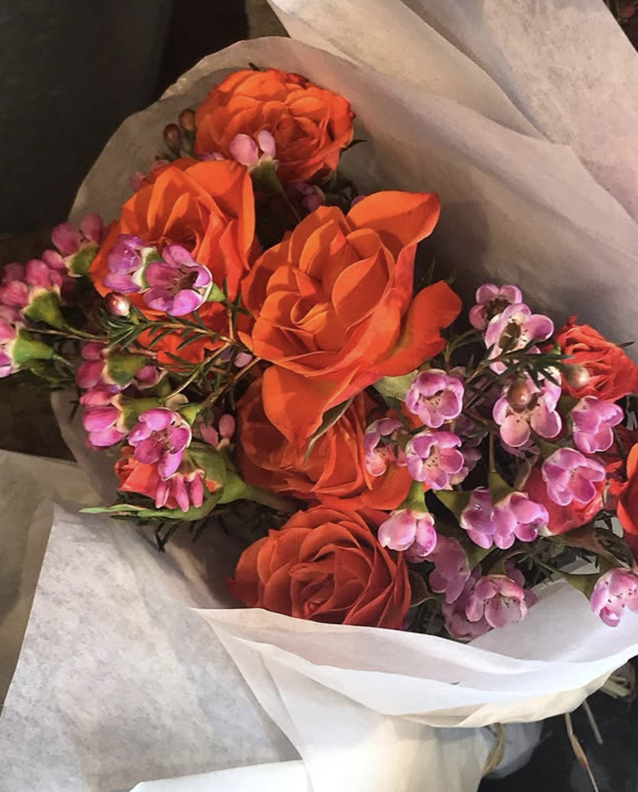 A bouquet of orange roses and pink wax flowers wrapped in white tissue paper.