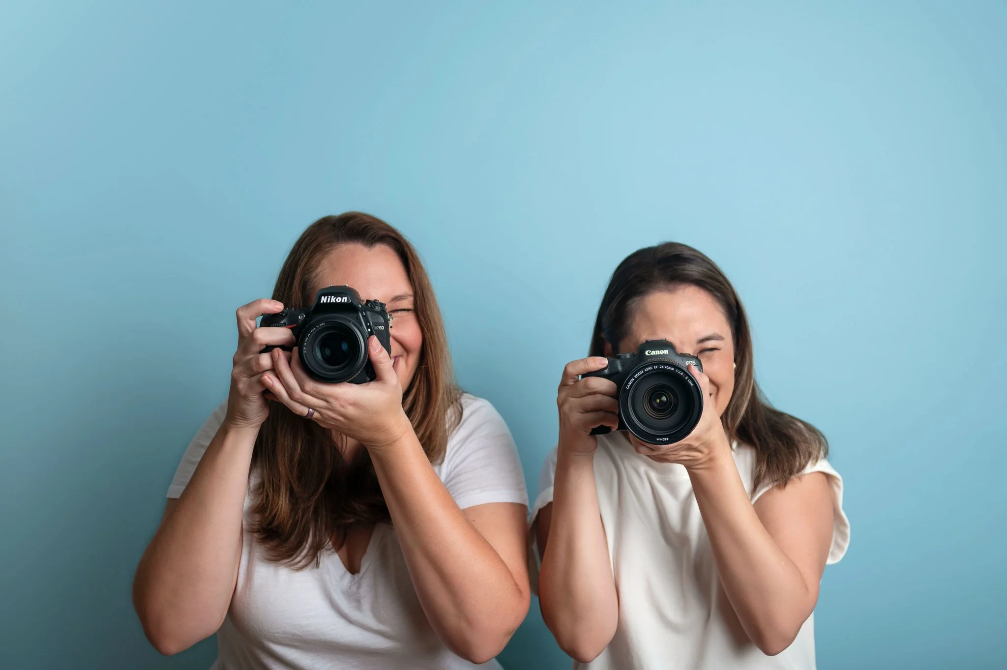 Two women smiling and taking photos with digital cameras against a light blue background.