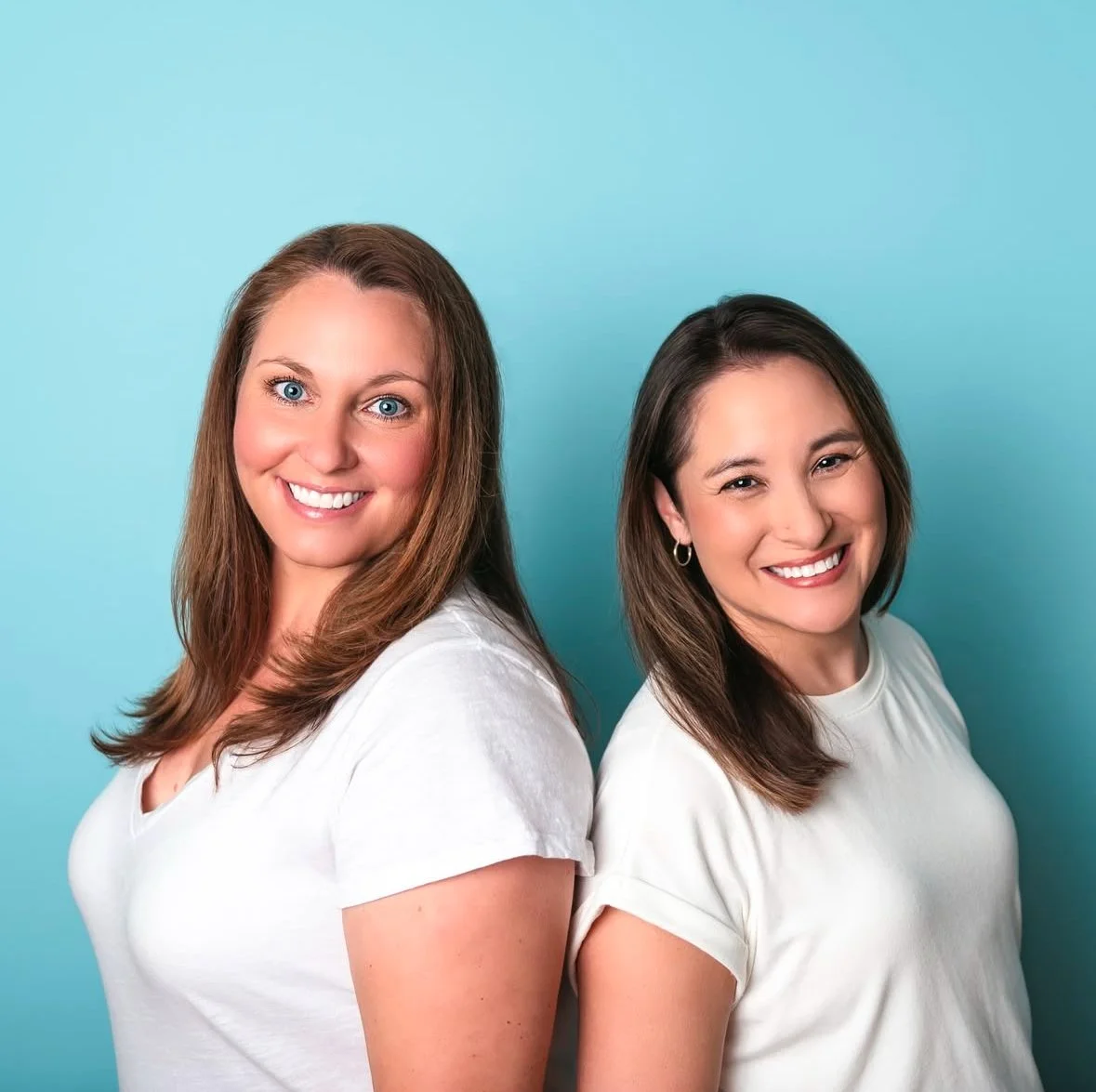 Two women smiling and standing back-to-back against a light blue background, wearing white t-shirts.