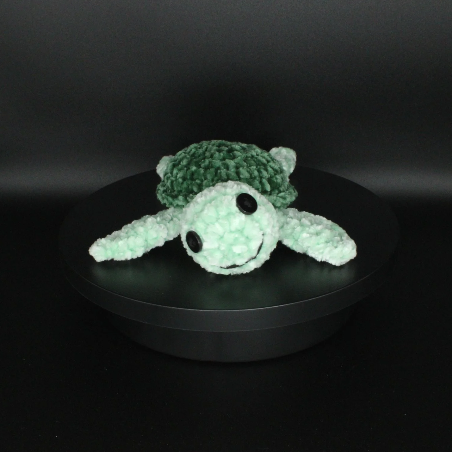 A plush toy resembling a turtle, with a green shell, white head and limbs, large black eyes, and a smiling face, placed on a black circular surface against a black background.