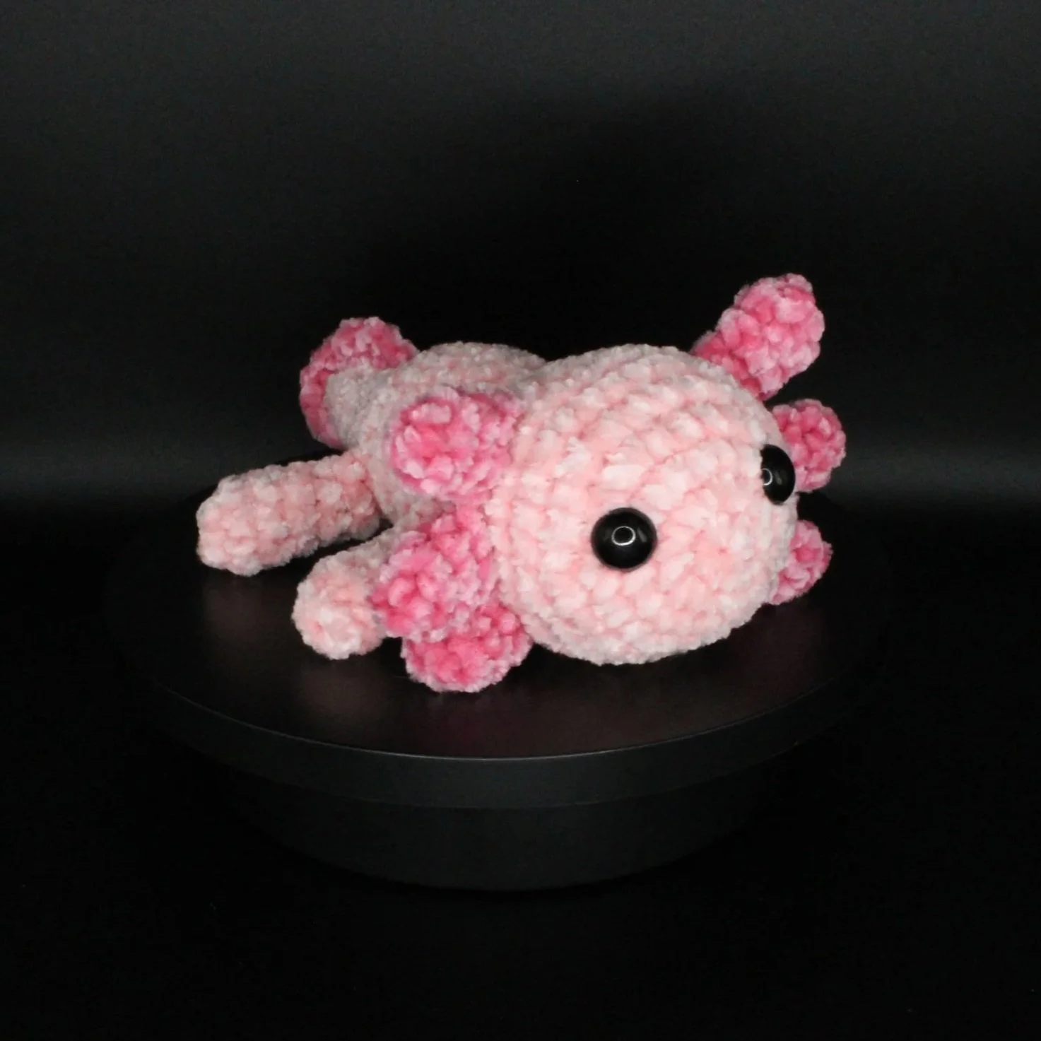 Pink plush toy crab with large black eyes, lying on a black surface against a black background.