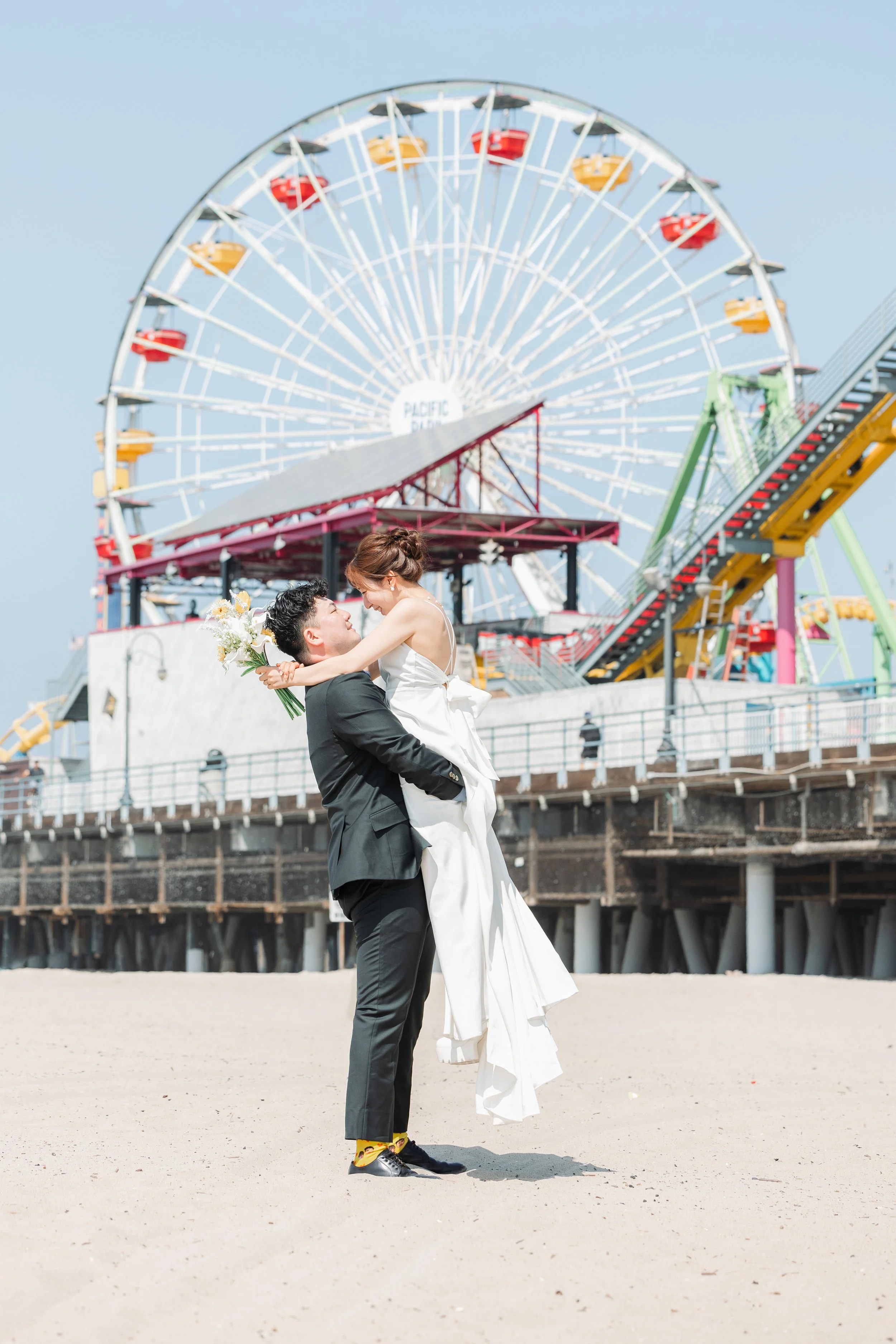 砂浜に立つ新郎が新婦を抱き寄せている。背景に観覧車と遊園地の乗り物が見える。