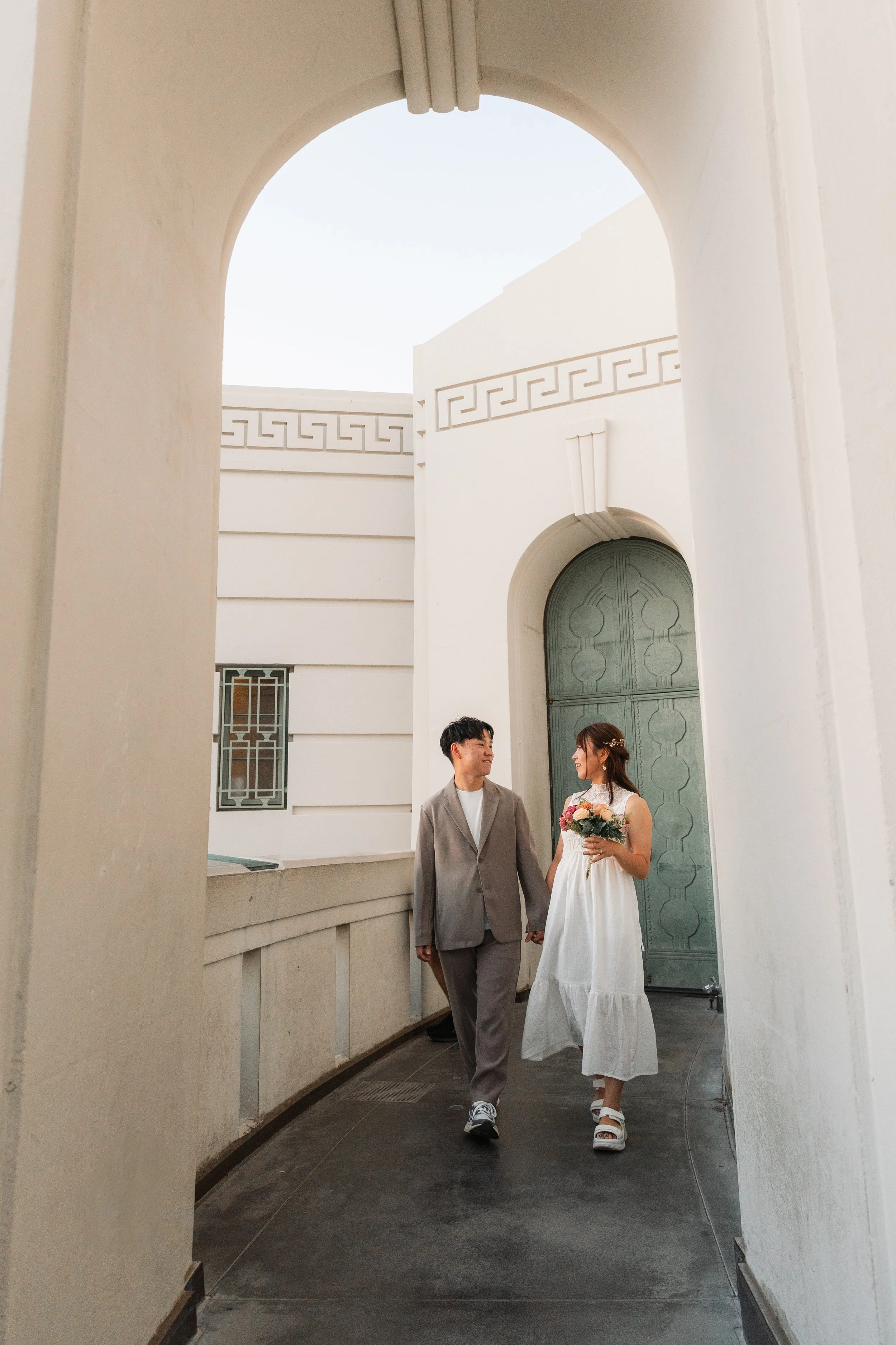 白い建物の通路を歩く若い夫婦。女性は花束を持ち、白いドレスを着ている。男性はグレーのスーツを着ている。