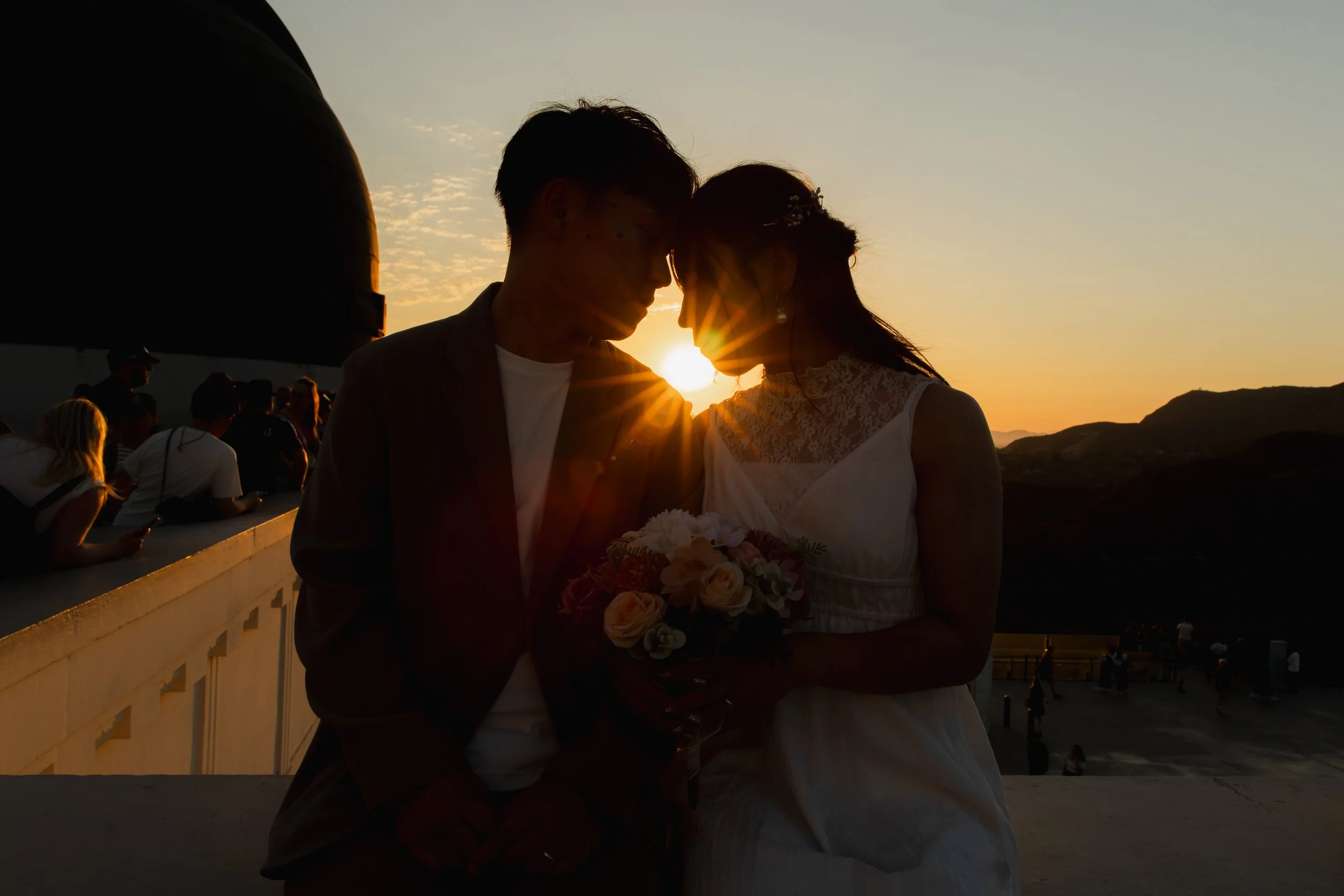夕焼けの中で寄り添う男女カップル。彼女は花束を持ち、二人は顔を近づけている。背景に観光客や山並みが見える。