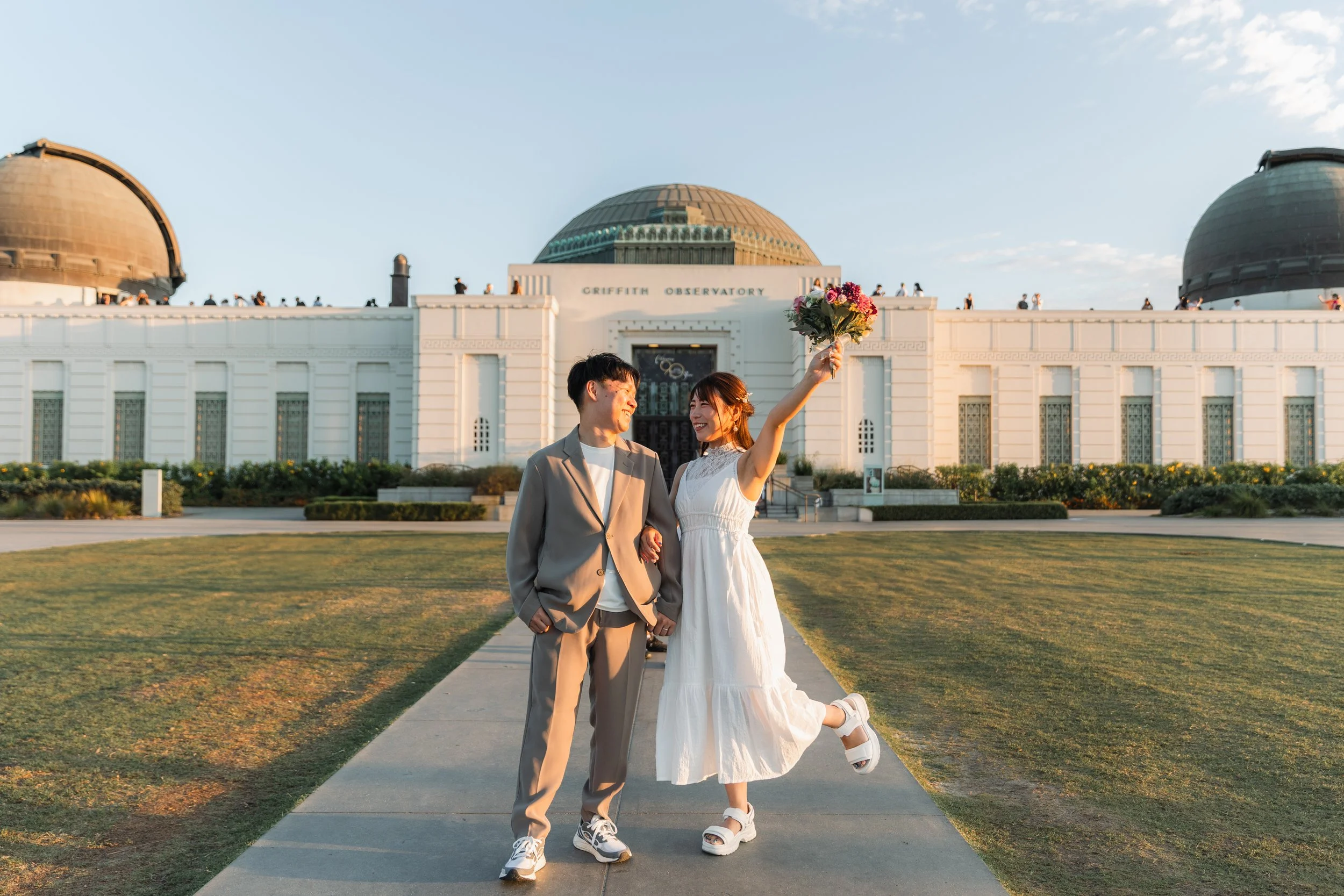 結婚式のカップルがグリフィス天文台の前で笑顔で手をつなぎ、花束を持ち上げるシーン