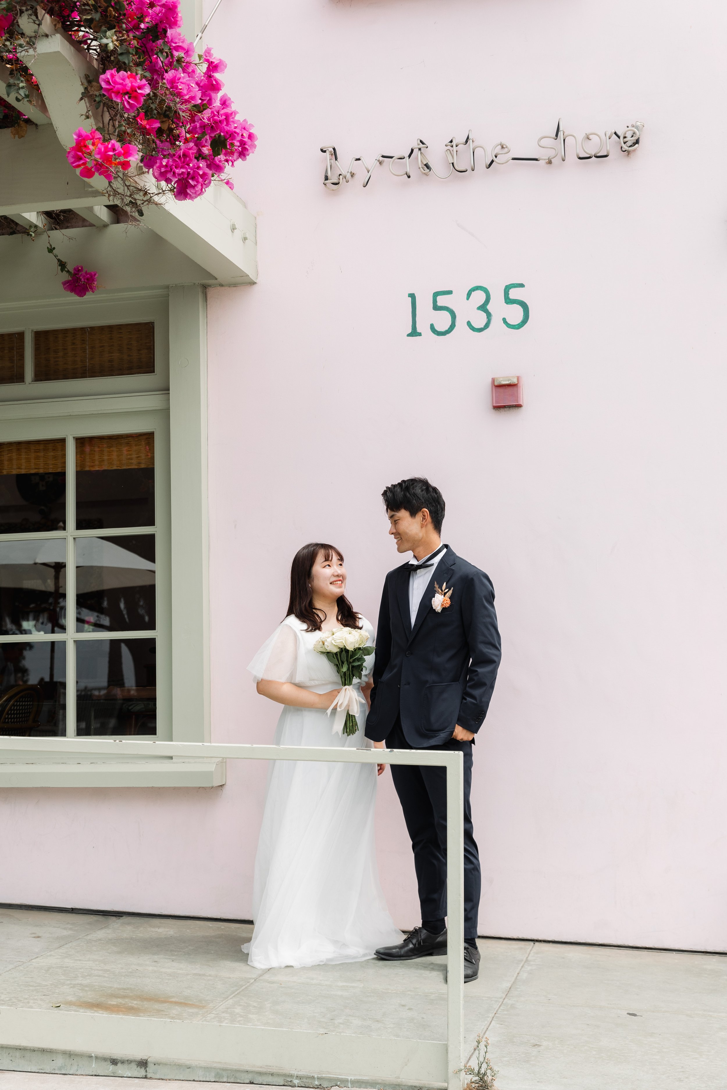 結婚式の新郎新婦が笑顔で見つめ合っている。女性は白いドレスを着て白い花束を持ち、男性はダークスーツを着ている。背景はピンクの壁に「Hydrate shore」と書かれ、住所の番号「1535」がある。