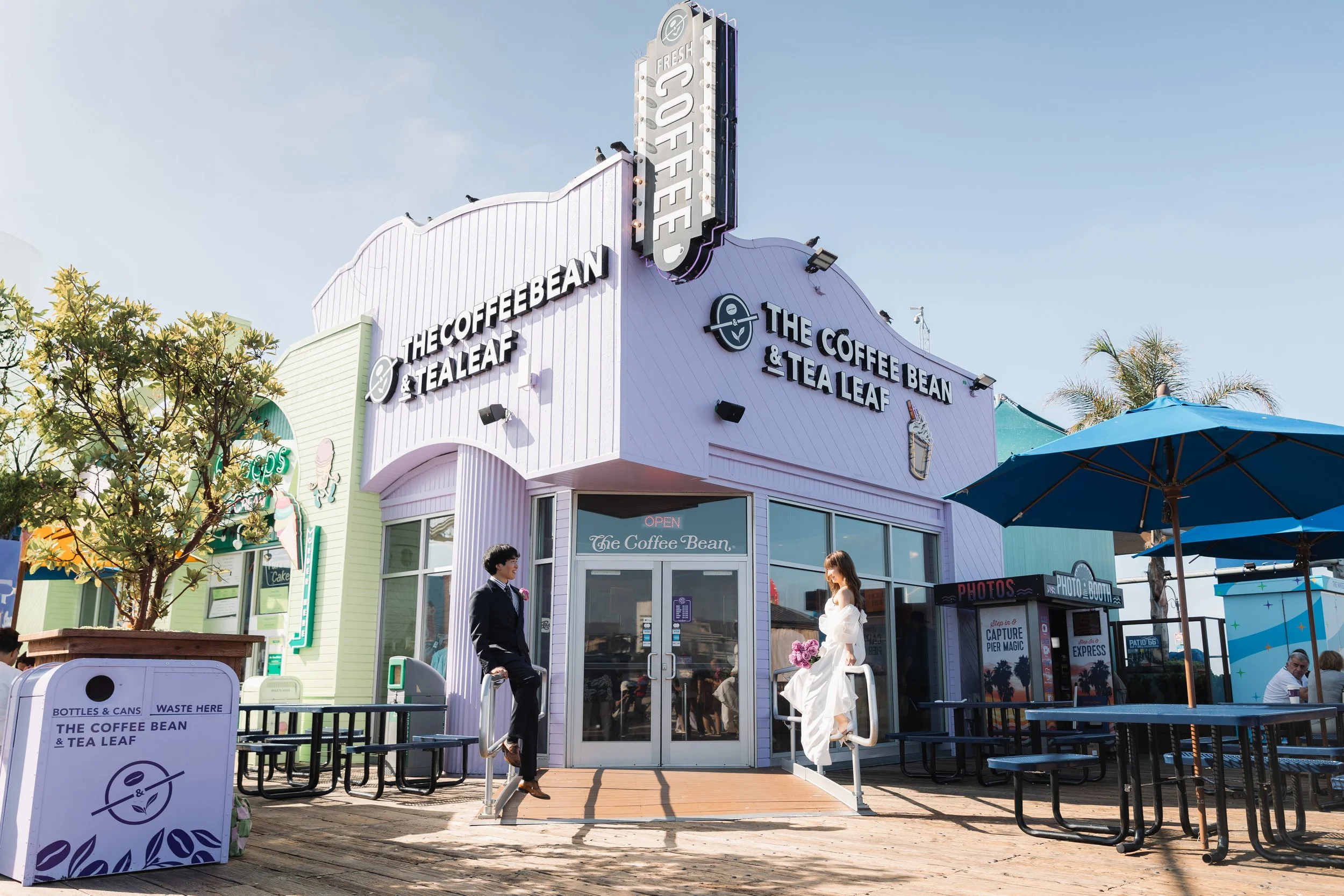 紫色の建物「THE COFFEE BEAN & TEA LEAF」と書かれた看板のカフェの前に男性と女性が座っている。青い日除けの下で屋台やテーブルと椅子が並ぶ。