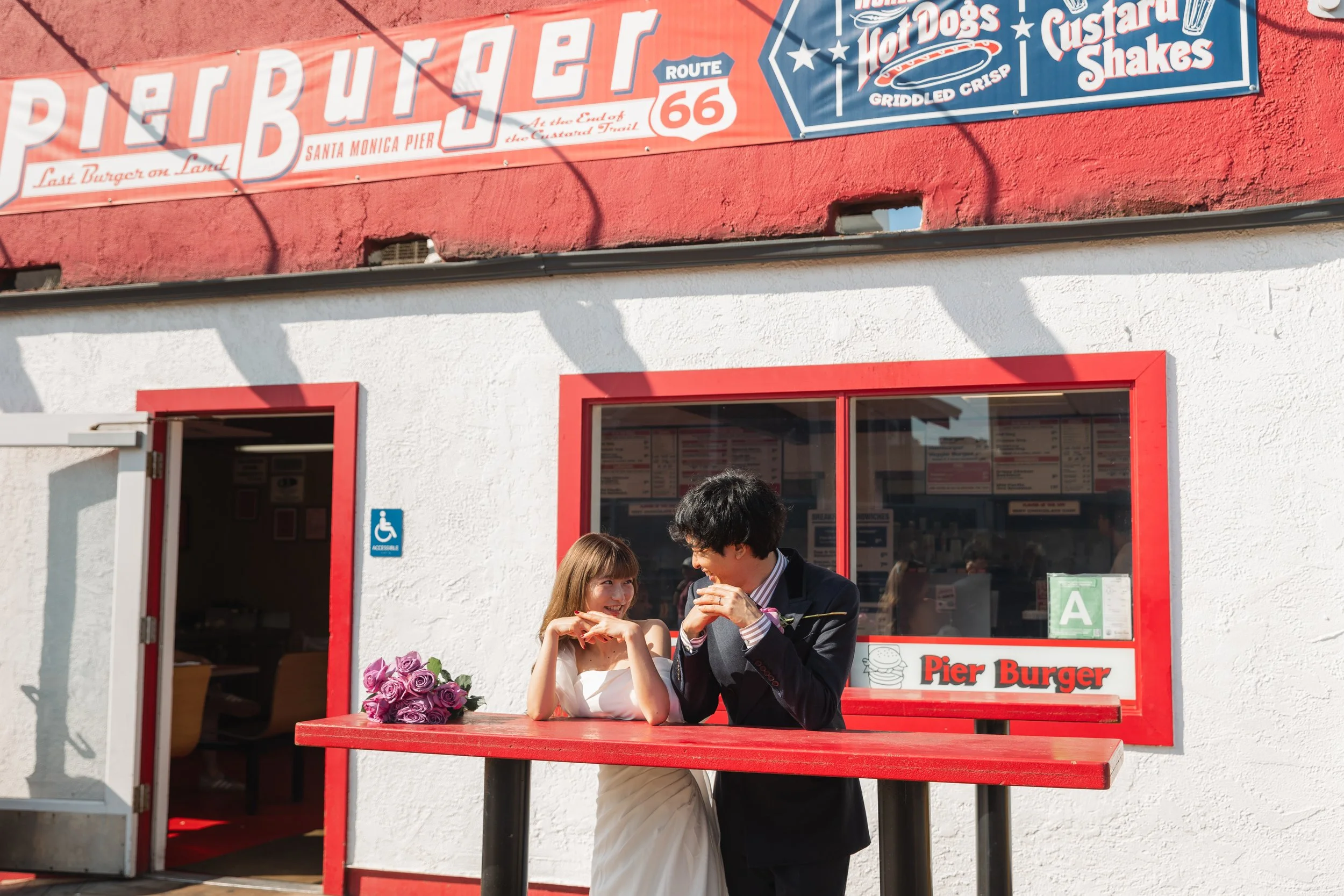 白い壁と赤い窓枠の建物前で、花束を持った女性と黒いスーツの男性が笑いながら交流している。