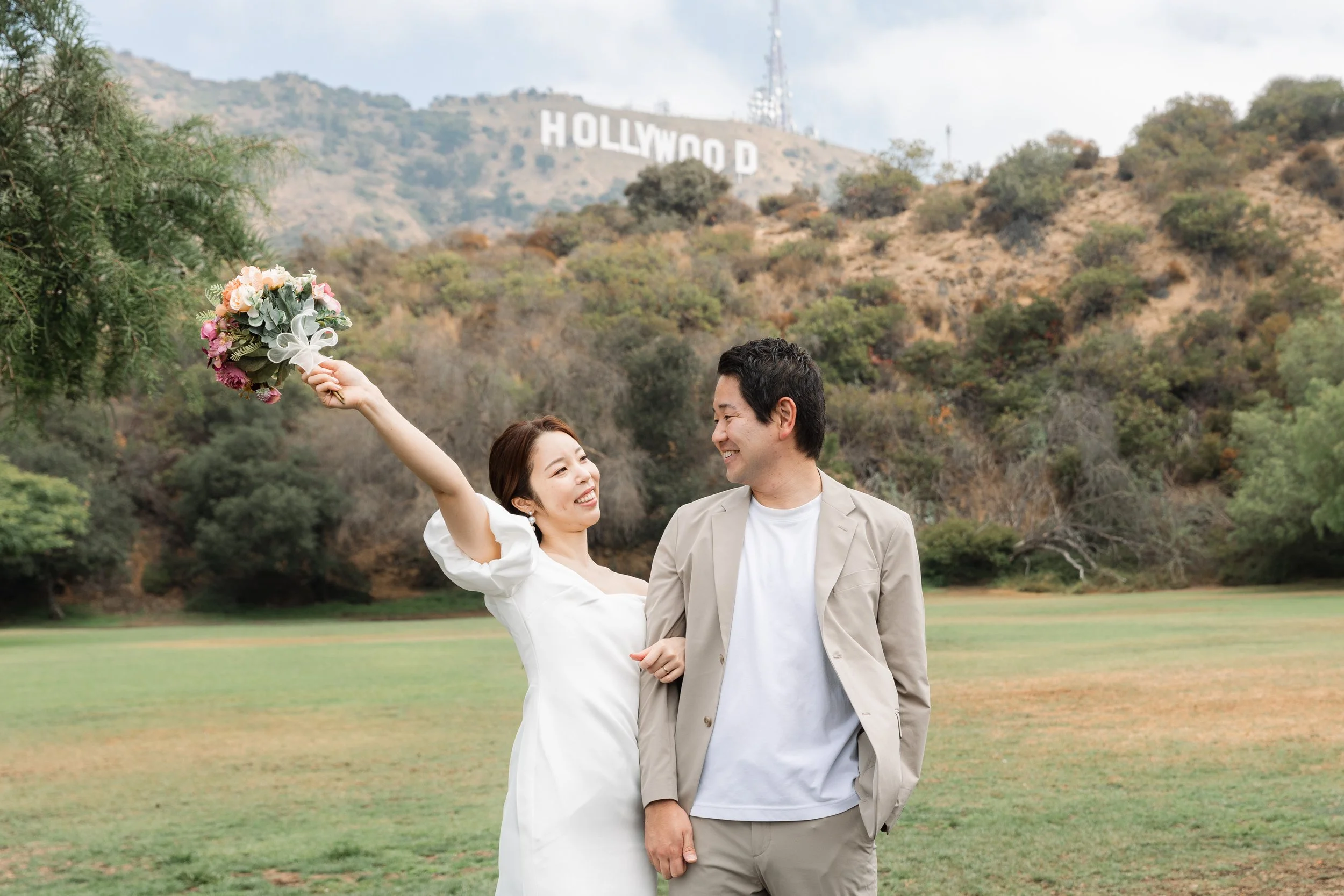 結婚式を挙げている新郎新婦のカップルが公園で楽しそうに笑い合っている。女性は花束を持ち、男性はピンクのシャツを着ている。背景にはハリウッドのサインが山の上に見える風景。