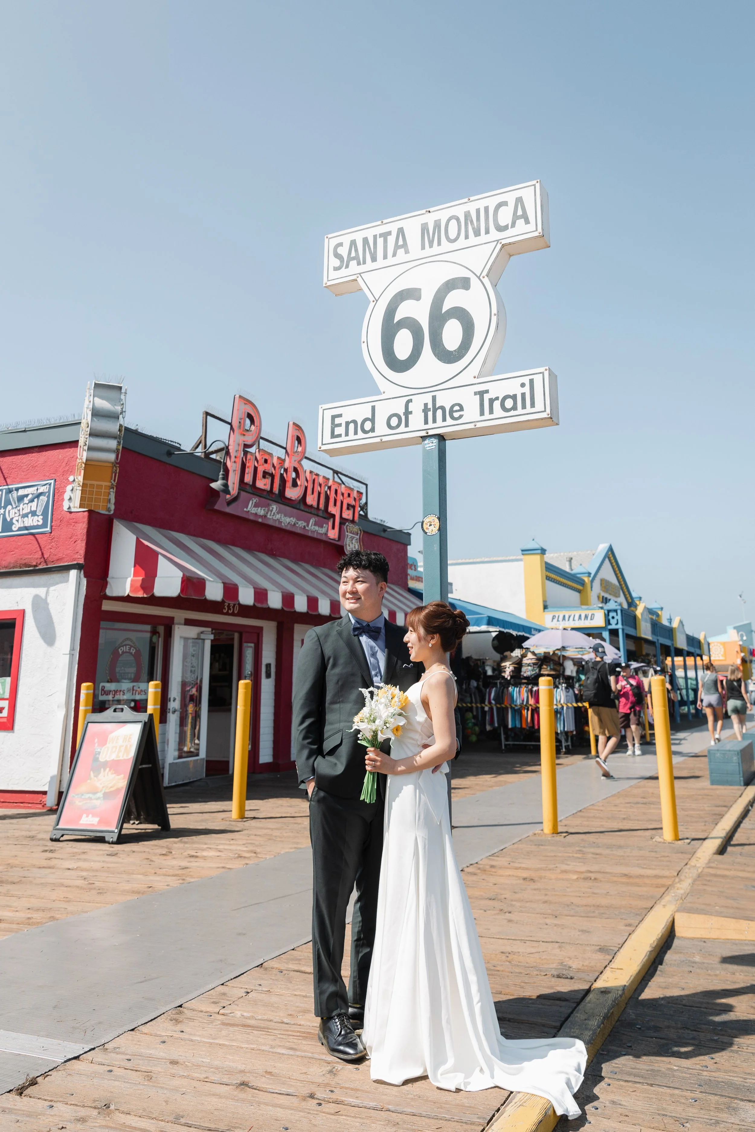 結婚した新郎新婦がサンタモニカの桟橋で写真を撮っている。背景にはレストランやショップが並び、空は青く晴れている。