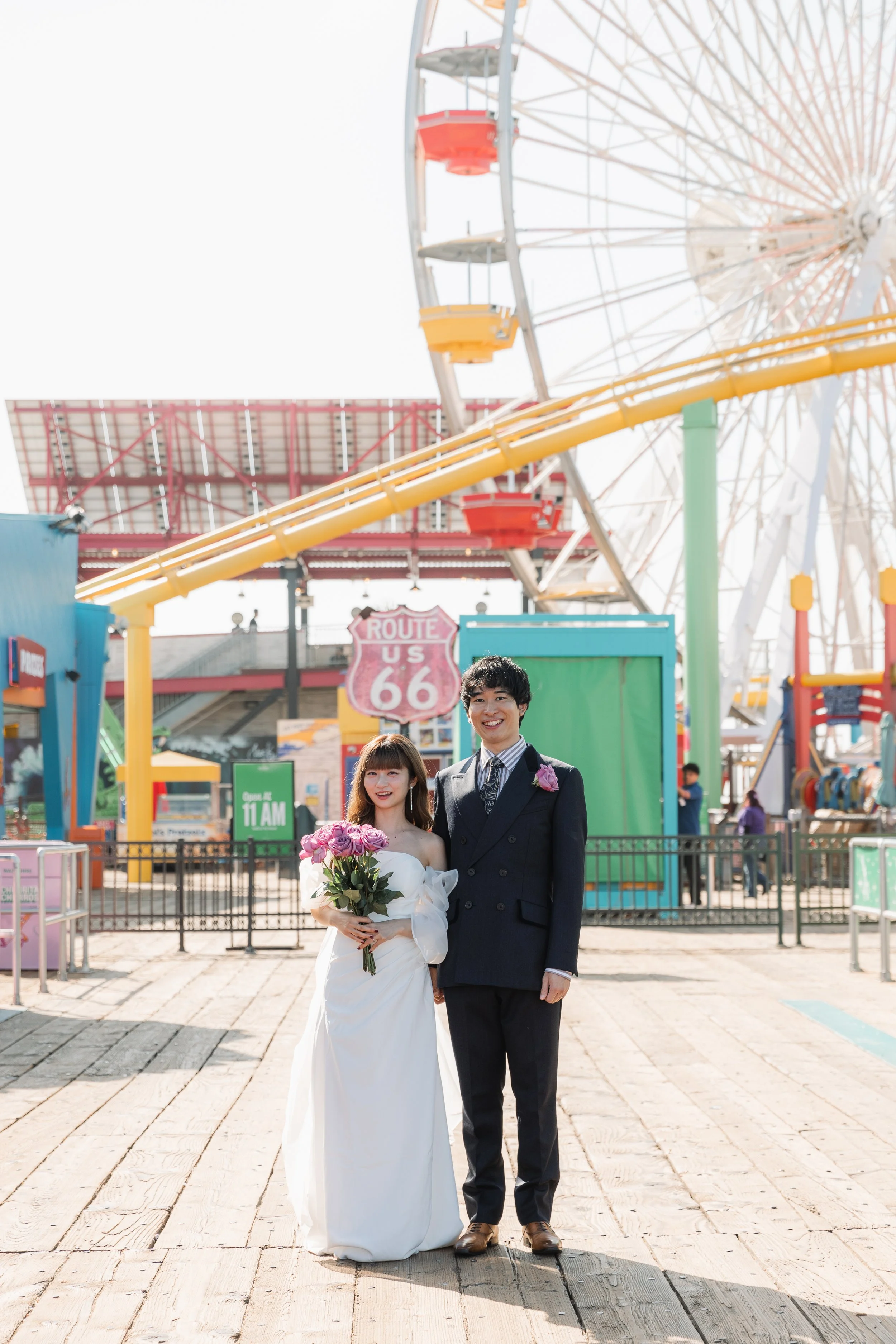 サンタモニカの遊園地で結婚式を挙げているカップルが写真に写っている。女性は白いドレスに花束を持ち、男性はスーツを着ている。背景には観覧車とジェットコースターが見える。