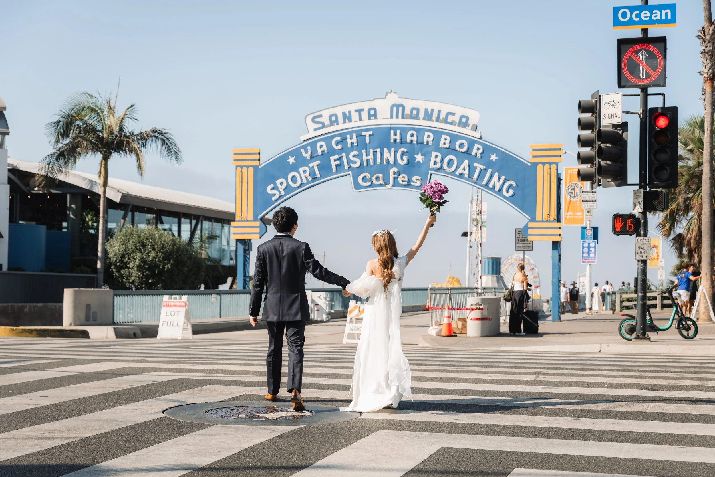 サンタモニカで結婚式を挙げるカップルが横断歩道を渡っている。女性は白いドレスを着て花を持ち、男性は黒いスーツを着ている。背景にはサンタモニカの看板と海が見える