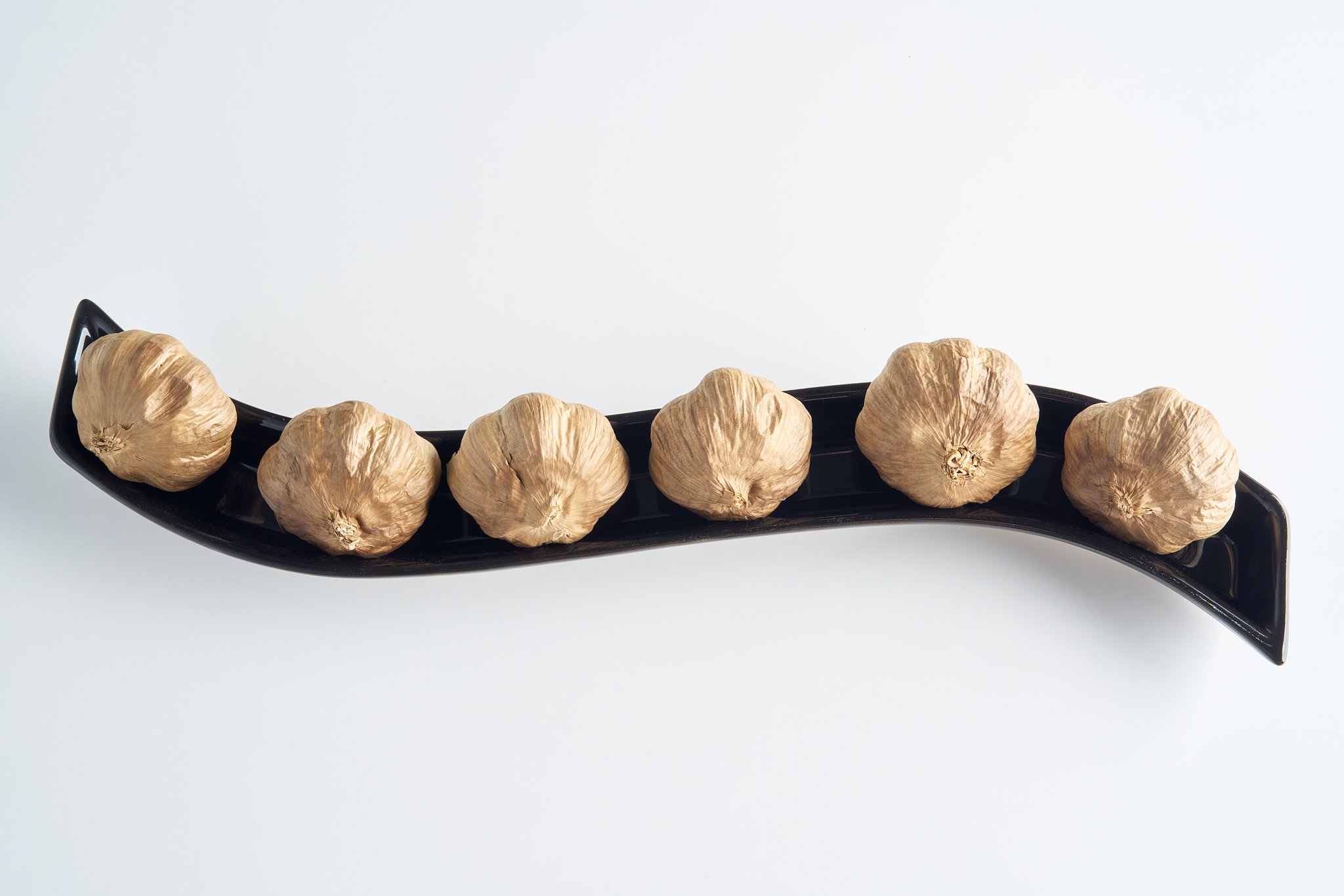 Six garlic bulbs on a curved black serving dish against a white background.