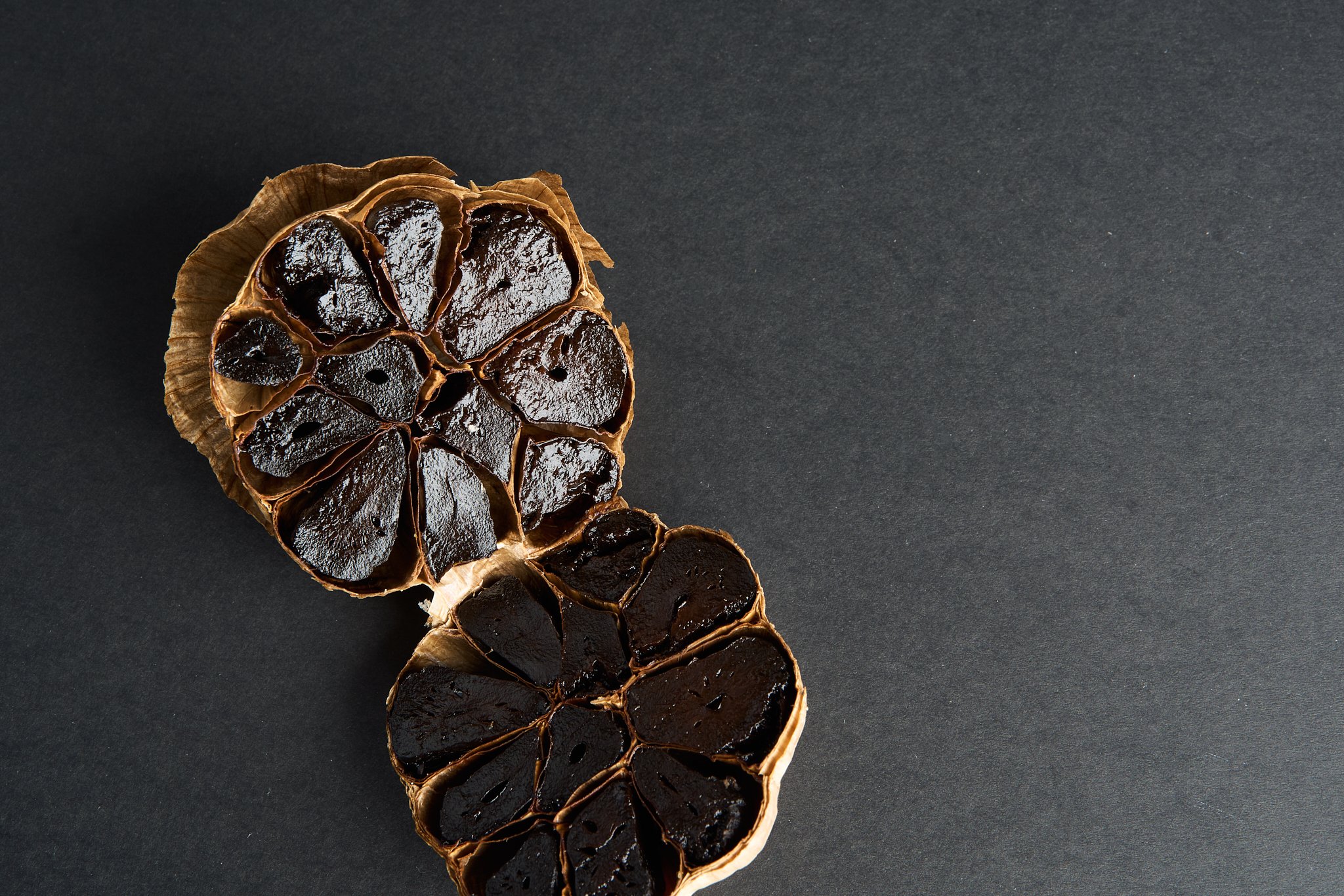 Two garlic bulbs with dark, roasted garlic cloves inside, placed on a dark textured surface.
