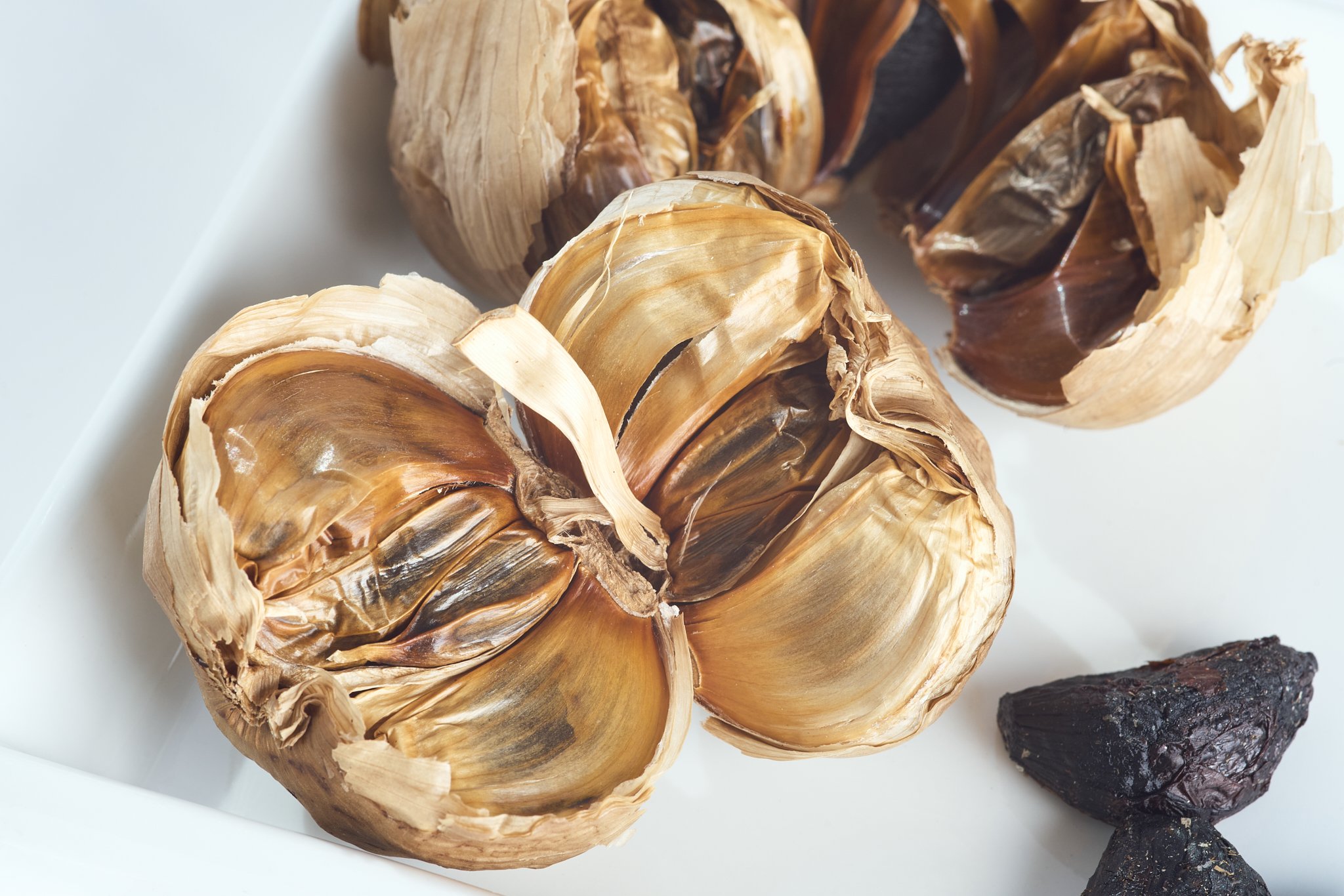 Close-up of roasted garlic bulbs on a white surface, with some cloves exposed, and a piece of black rock at the bottom right.