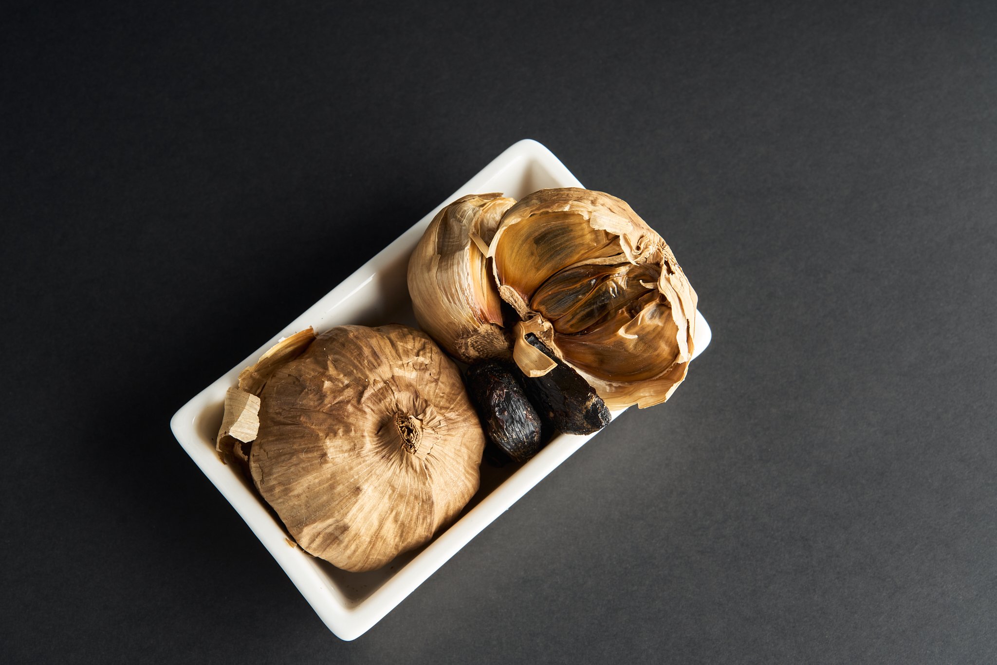 A small white rectangular dish containing garlic bulbs and black olives on a dark surface.