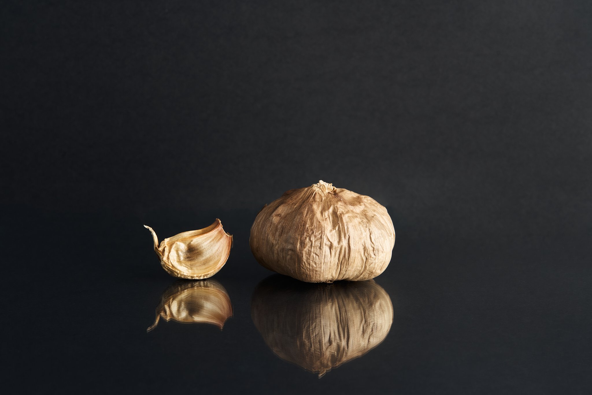 Whole garlic bulb with one garlic clove beside it on a reflective black surface against a dark background.