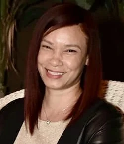 Portrait of a woman with shoulder-length auburn hair, smiling, wearing a black jacket and a light-colored top, sitting indoors with green plants in the background.