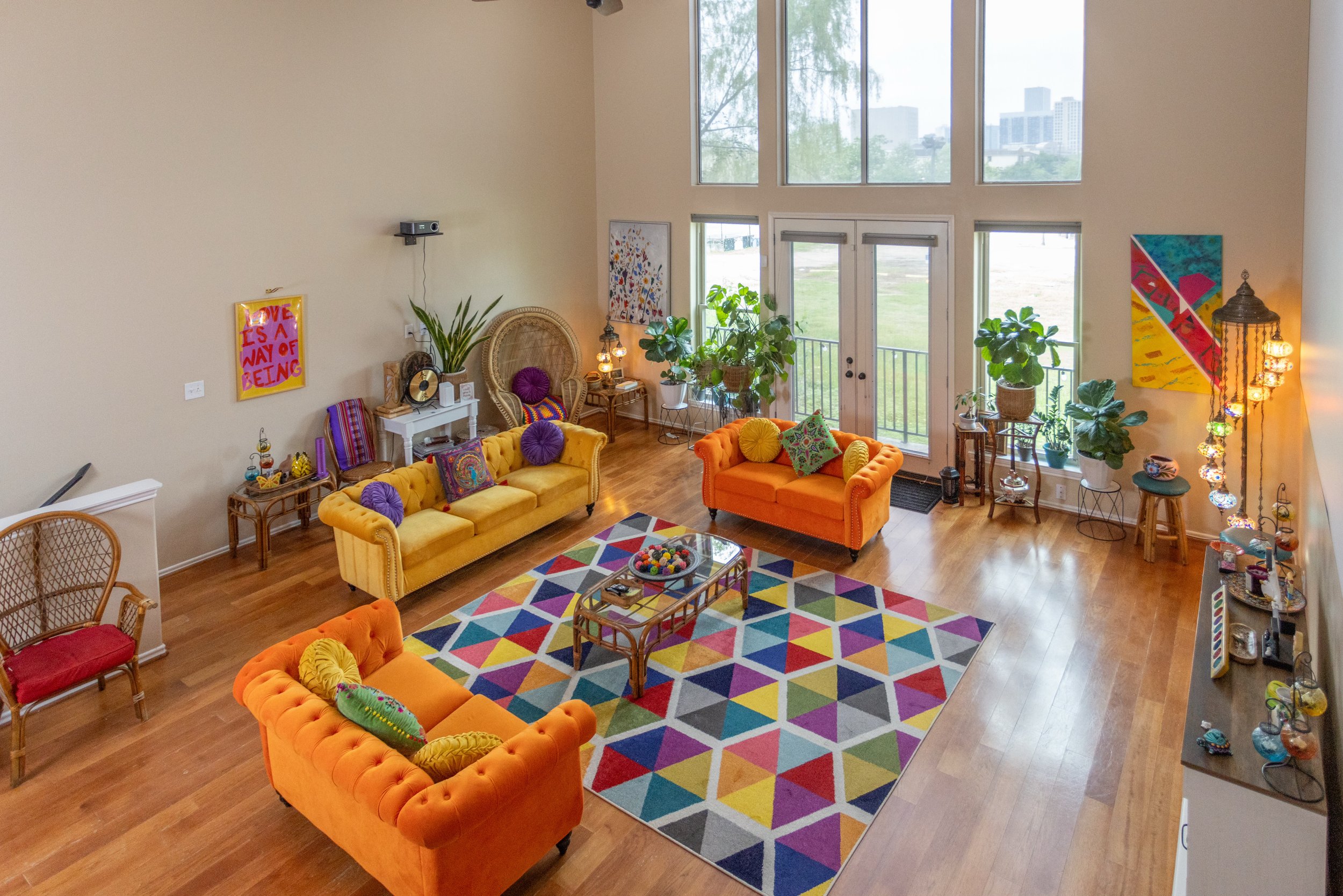 Brightly colored living room with orange and yellow sofas, a geometric patterned rug, large windows, and indoor plants.