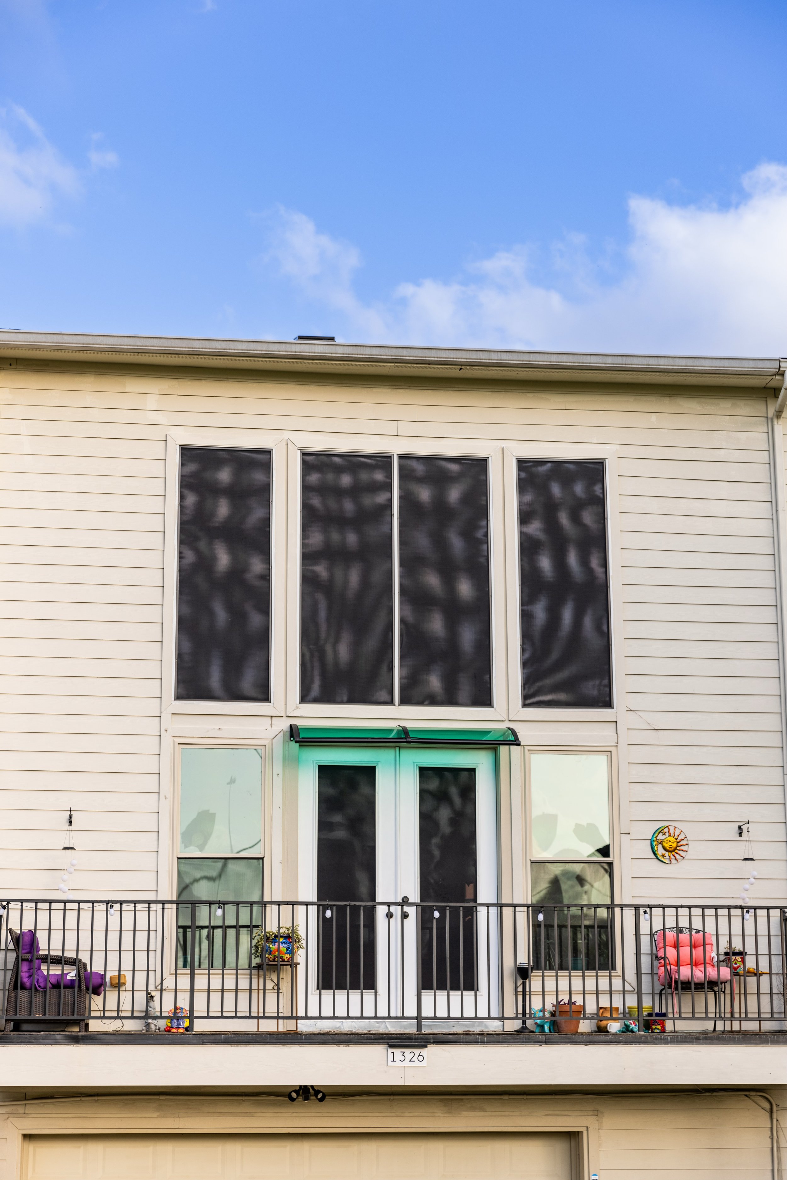 Exterior view of a house featuring large windows with screens, a balcony with a railing, and patio furniture. Decorative plants and a sun wall art are visible on the balcony. The house number '1326' is displayed above a garage door. The sky is blue w