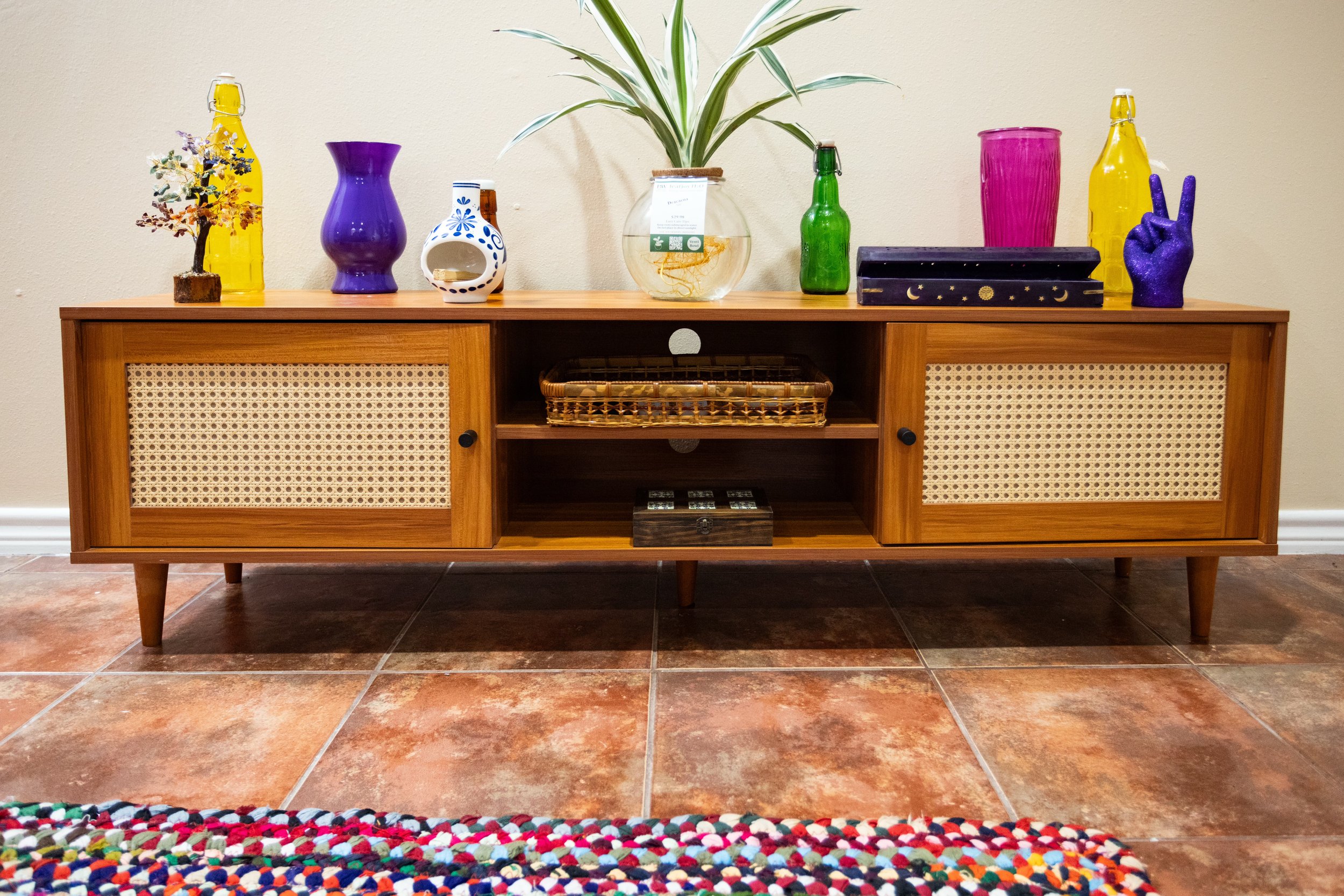 Mid-century modern wooden cabinet with rattan doors displaying colorful vases, bottles, a potted plant, and decorative items on top, situated on a tiled floor with a multicolored rug.