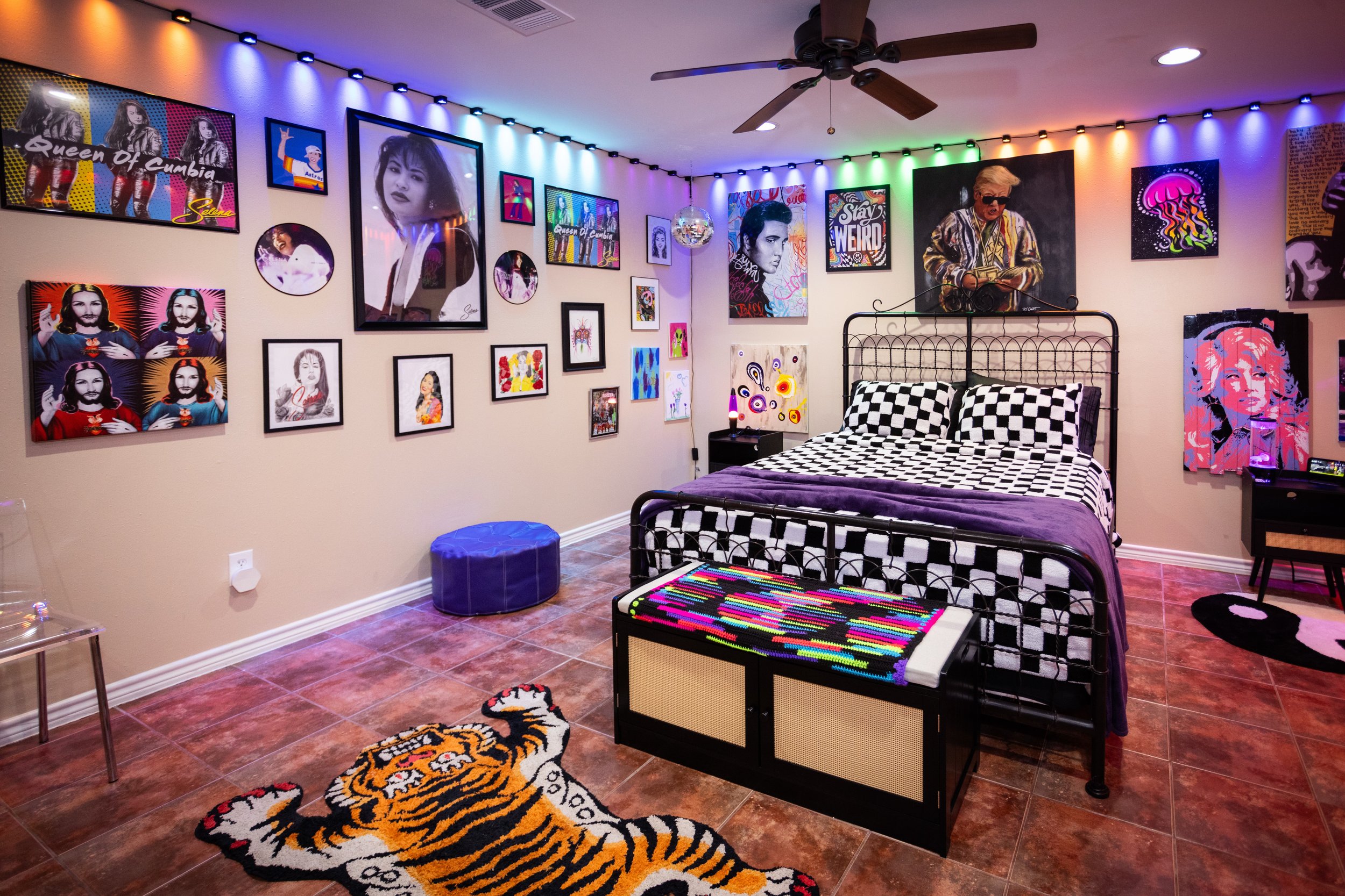 A vibrant bedroom with colorful pop art and music-themed wall decorations, a checkered bedspread, a ceiling fan, string lights, a clear chair, a blue ottoman, and a tiger rug on tiled flooring.