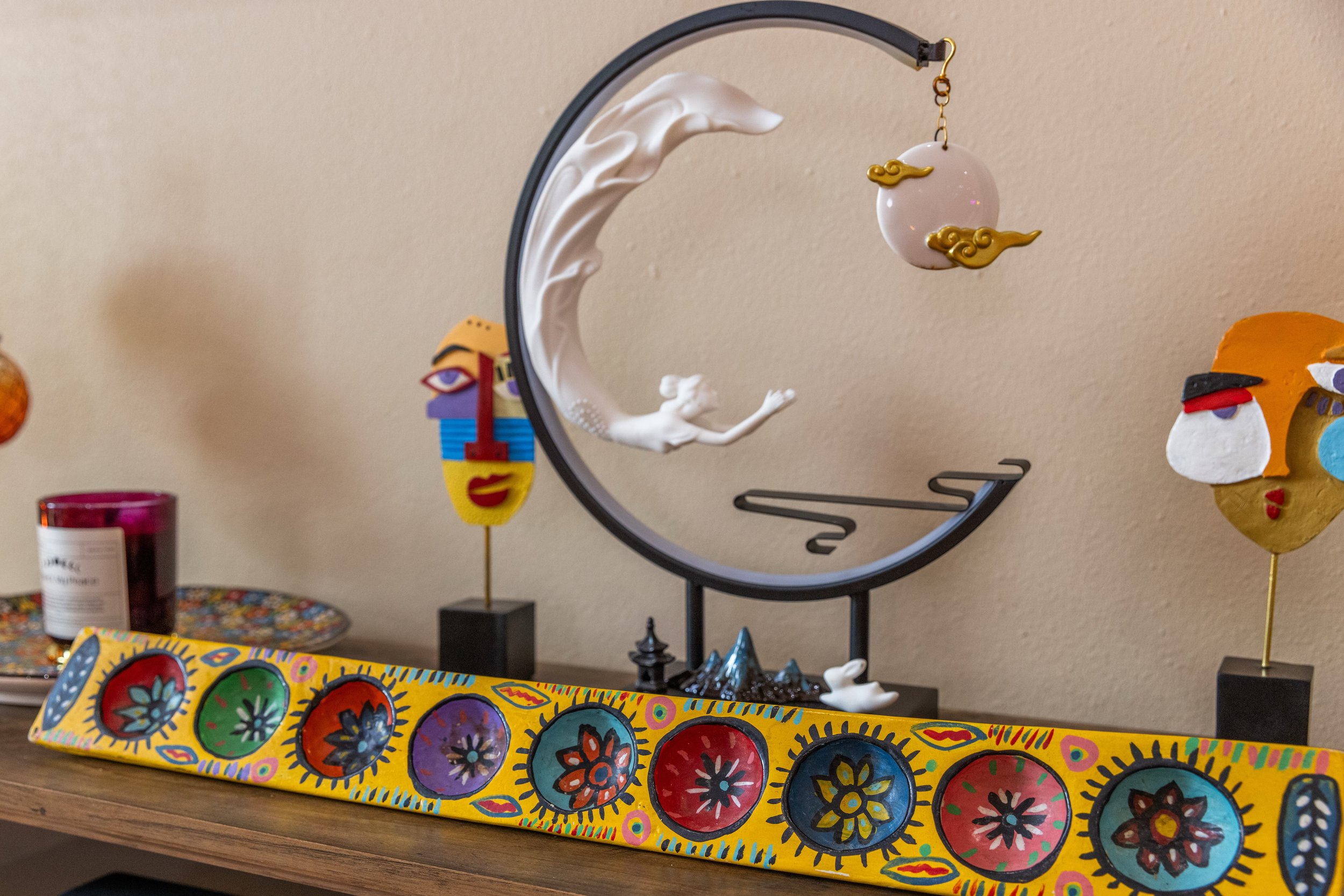 A decorative arrangement featuring a white figurine reaching towards a hanging ornament in a circular frame, colorful abstract masks on stands, a vibrant painted wooden plank with floral designs, and a candle in a glass jar on a wooden shelf.