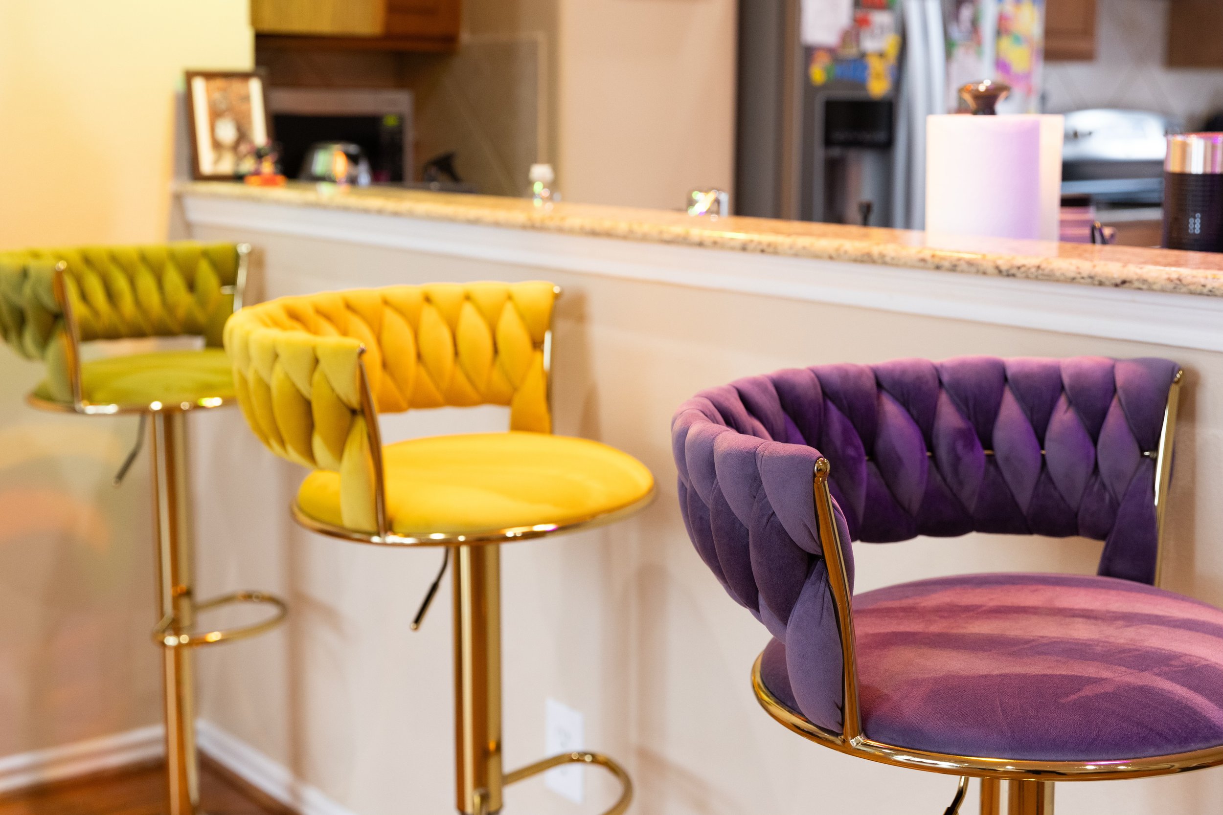 Colorful bar stools with plush cushioned seats and backs in purple, yellow, and green arranged along a kitchen counter.