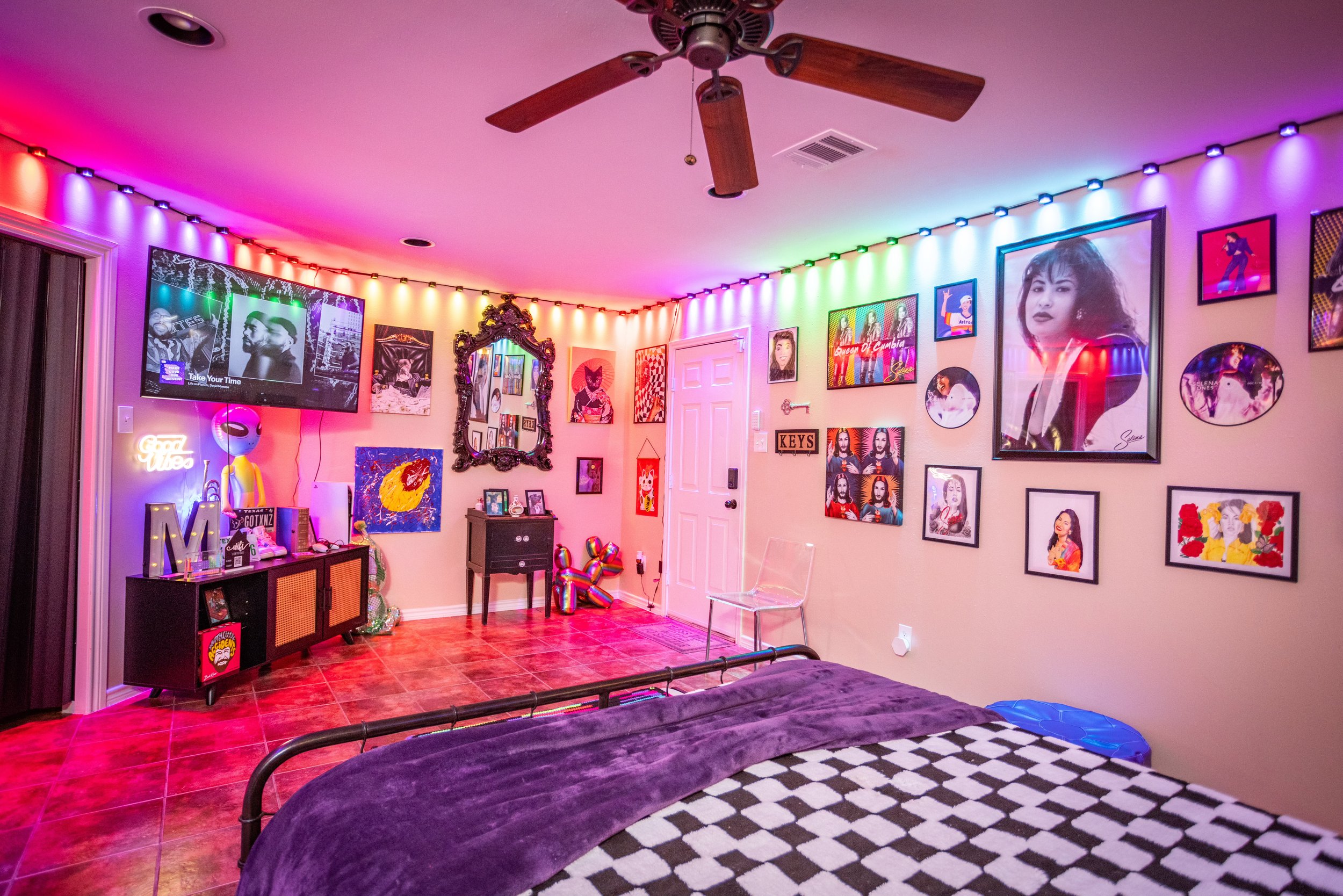 A colorful room with LED lights on the ceiling, artwork covering the walls, and a modern ceiling fan. There's a bed with a checkered blanket and a purple throw. A flat-screen TV is on, and a console table has decorative items. The room has tile floor