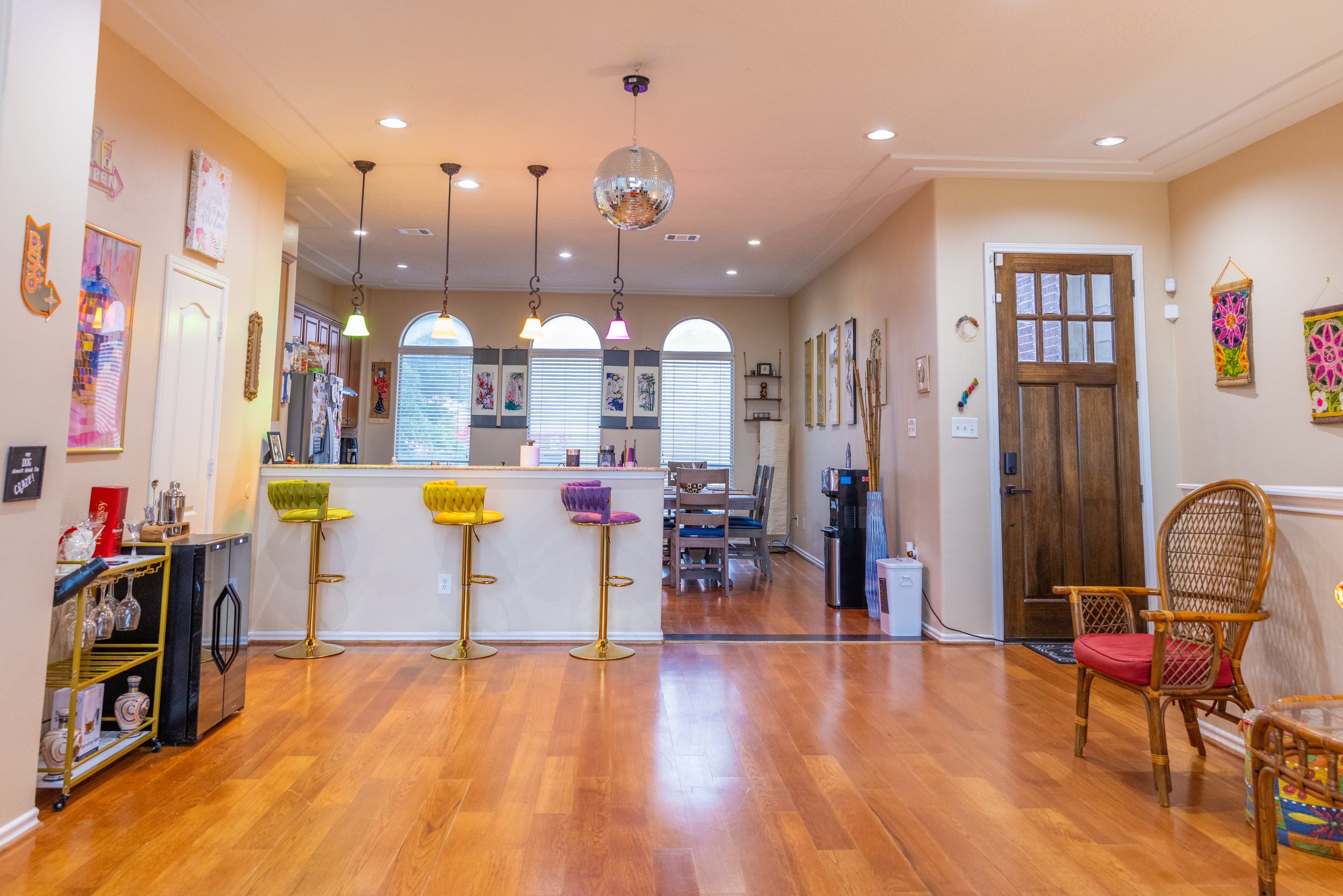 Spacious kitchen and dining area with wooden floors and eclectic decor, featuring a bar with colorful stools, pendant lights, and a disco ball. The dining table is visible in the background, alongside various wall art and an open entrance door.