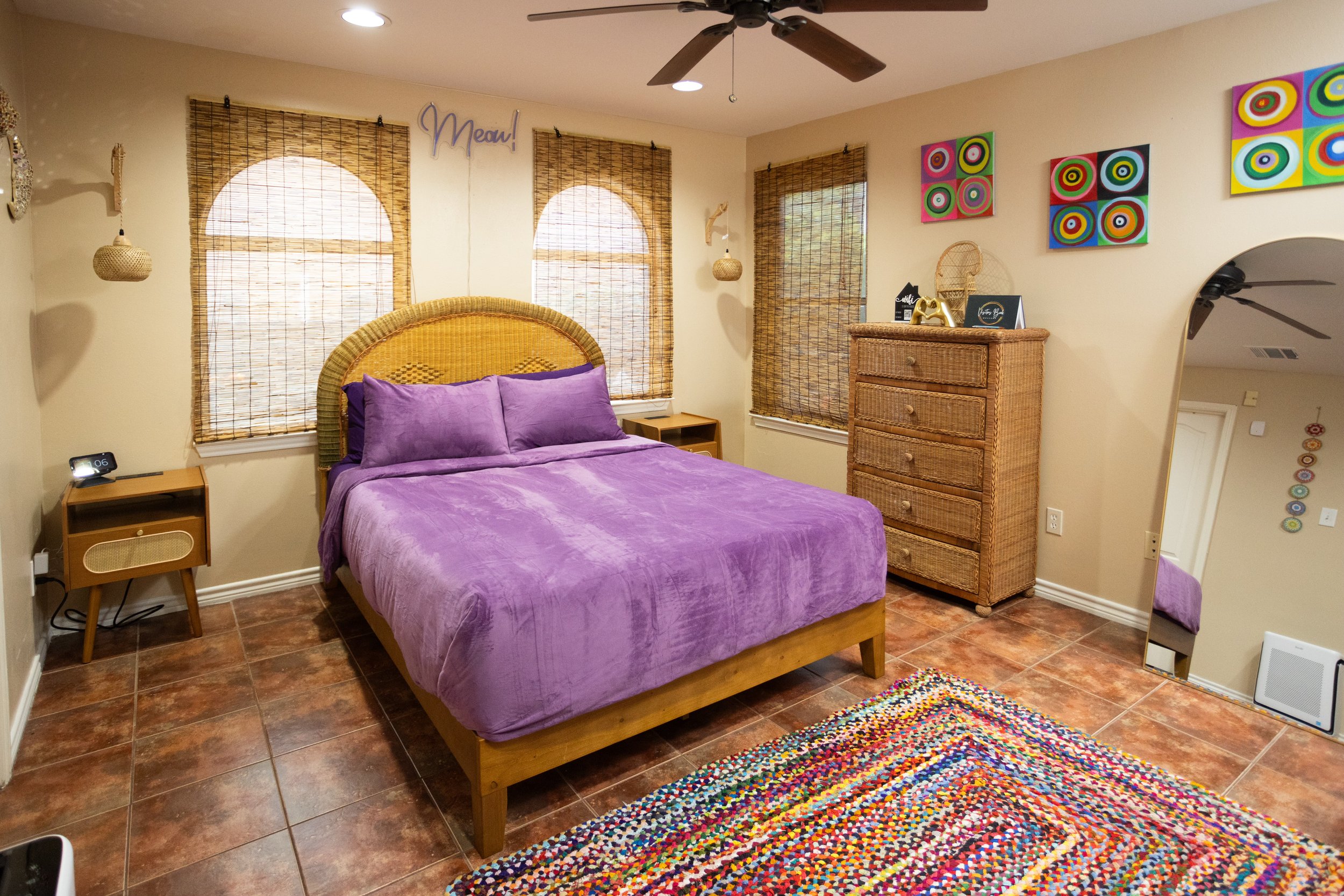 Bedroom with a purple bedspread, wicker furniture, colorful wall art, and a multicolored rug on tiled floor.