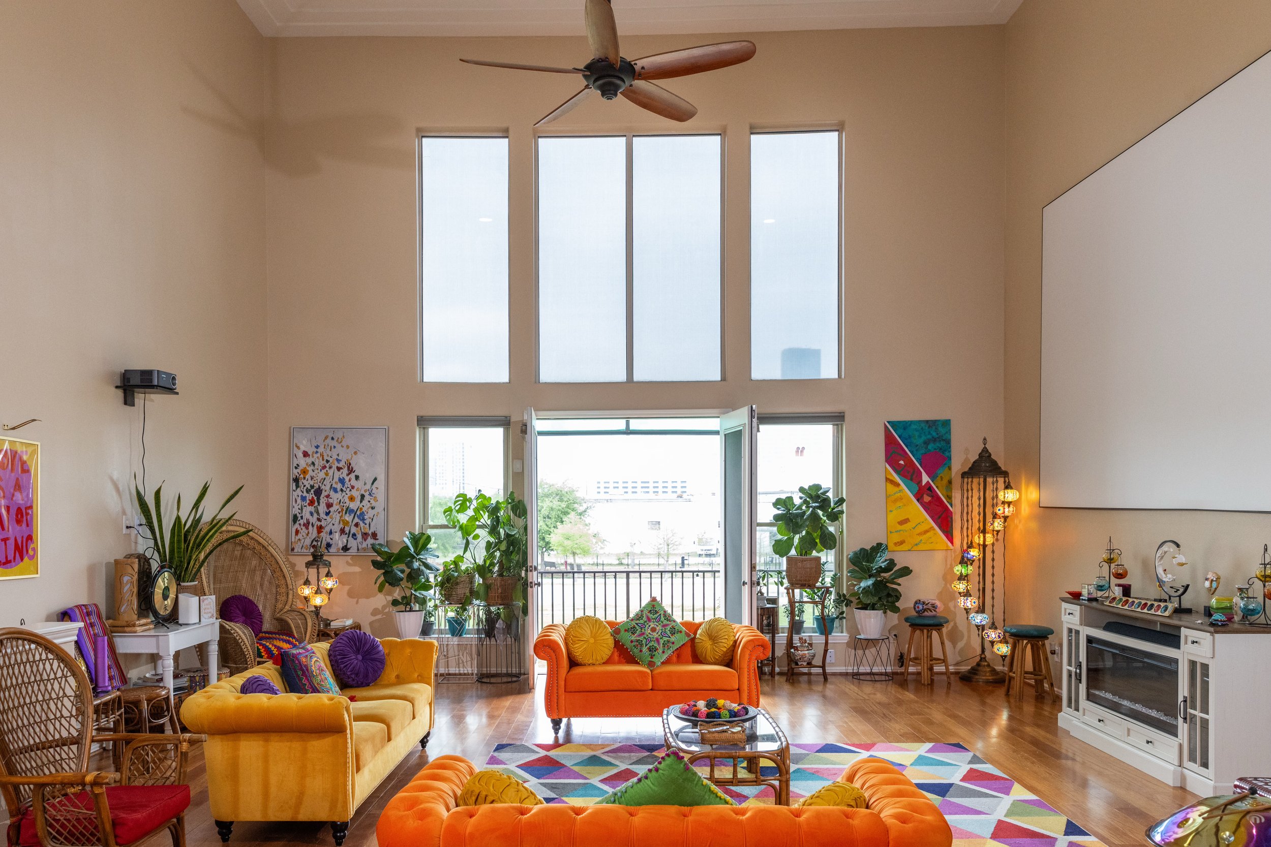Bright, colorful living room with high ceiling, large windows, orange and yellow couches, geometric rug, and indoor plants.
