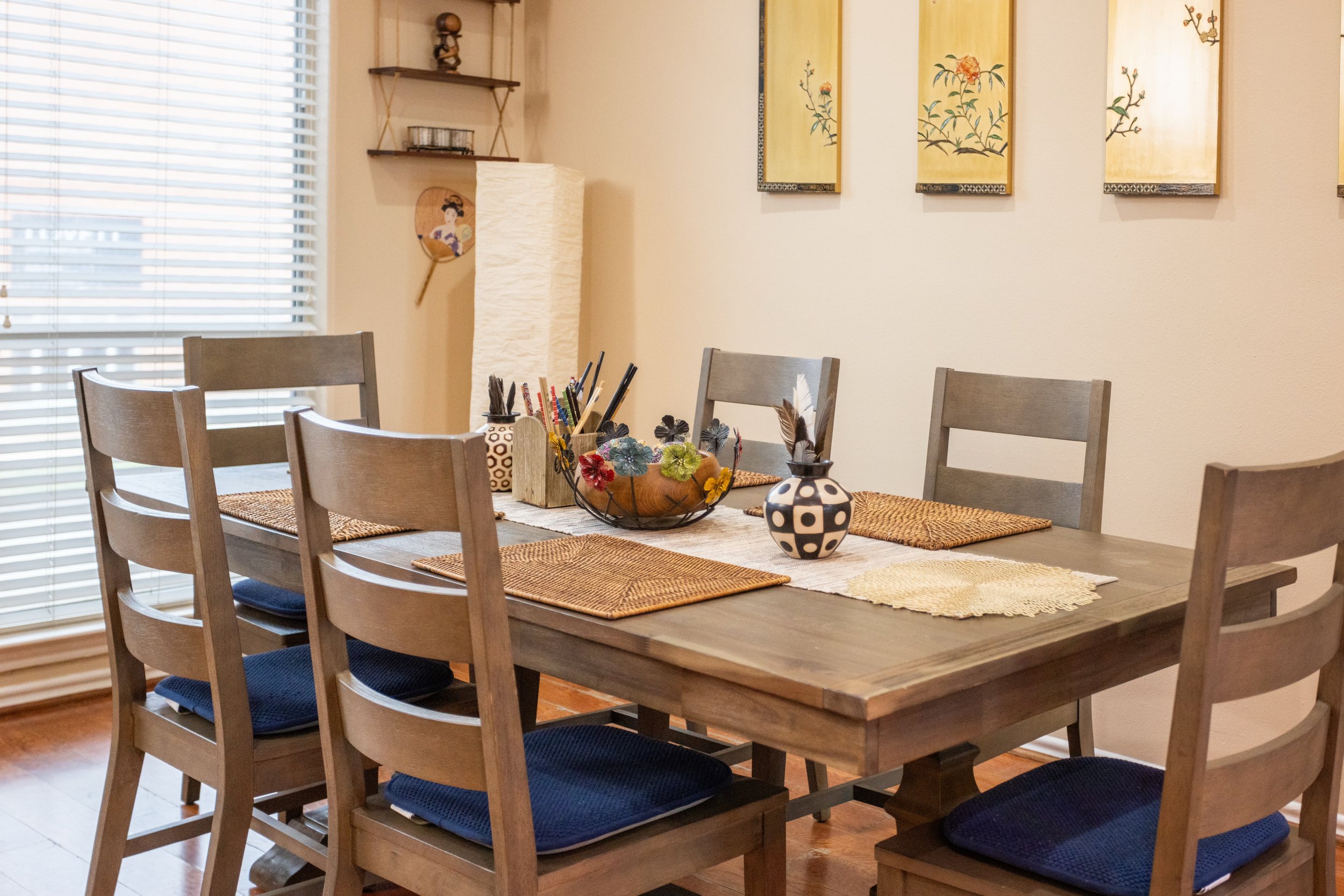 Dining room with wooden table and chairs, decorative placemats, centerpiece with colorful flowers, and wall art. Blinds cover a window, and a shelf holds decor items.