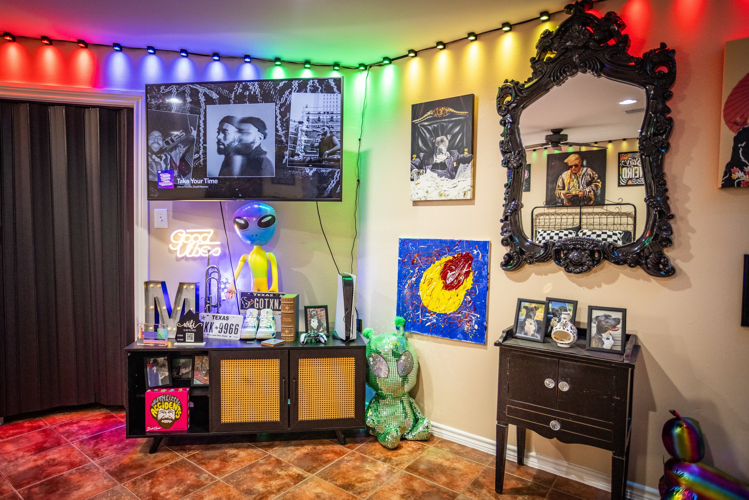 Colorful room with LED lights, wall art, a TV displaying an album cover, decorative mirrors, and alien figures. Includes framed photos, a cabinet with items like a neon sign, Texas license plate, and art pieces.