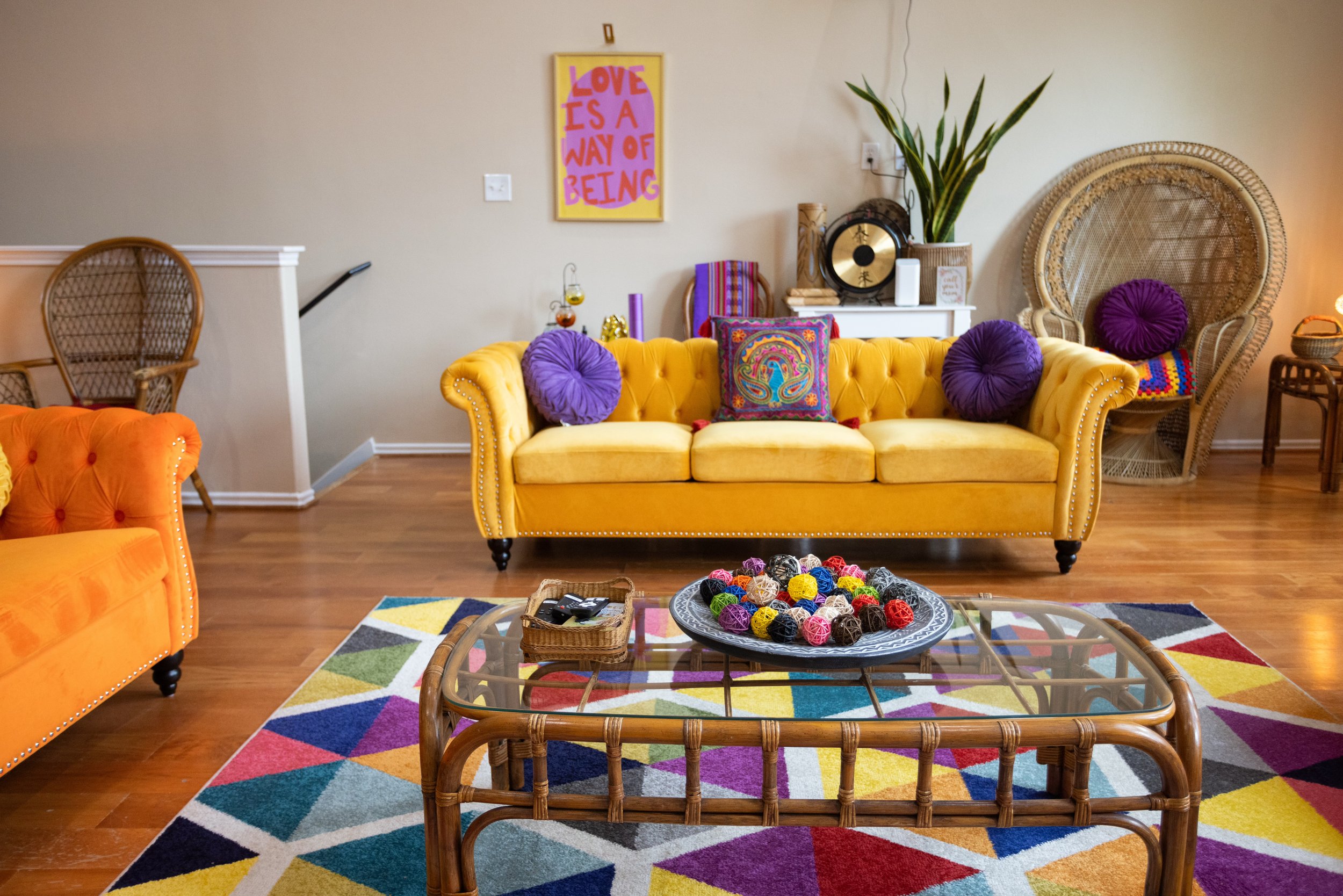 Colorful living room with yellow and orange sofas, purple accent pillows, a decorative geometric rug, and a glass-top wicker table. Wall features a motivational sign, "Love is a Way of Being."