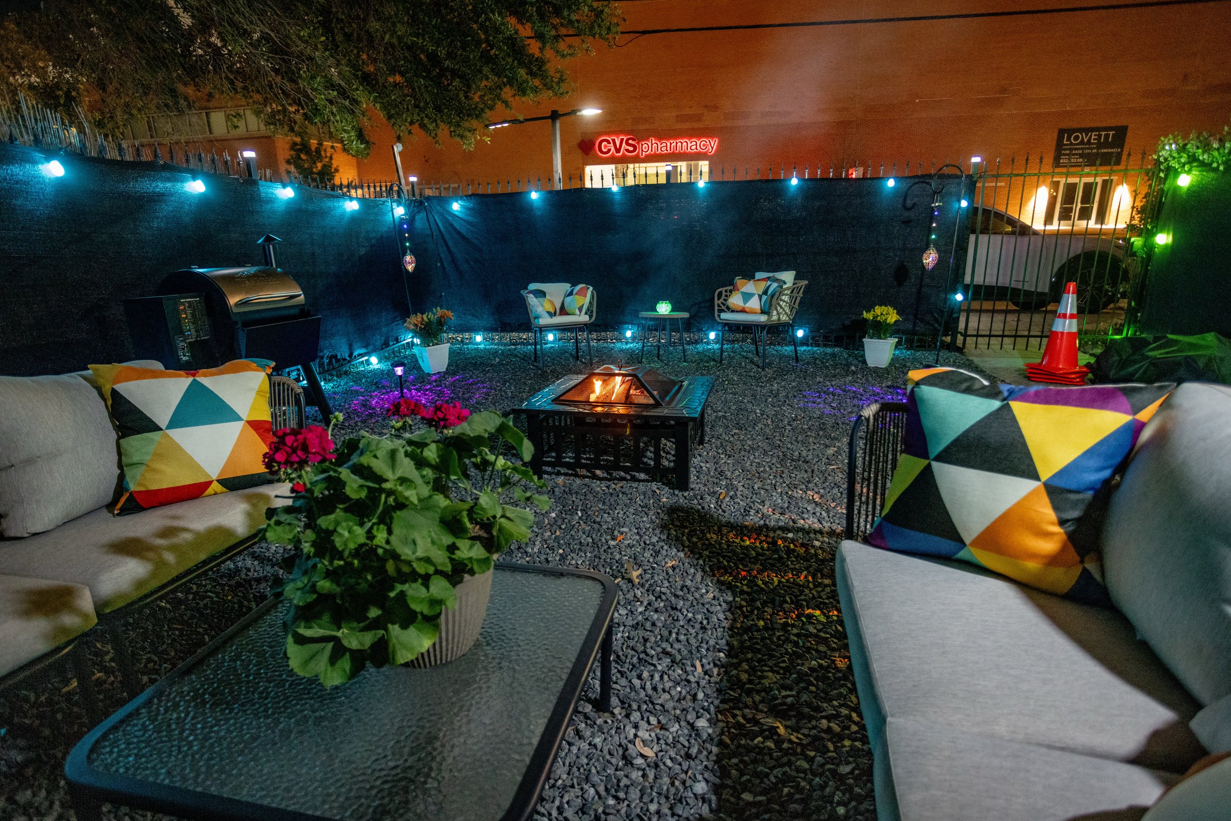Outdoor patio area with modern furniture, colorful geometric pillows, decorative lighting, fire pit, and potted plants; CVS Pharmacy sign visible in the background.