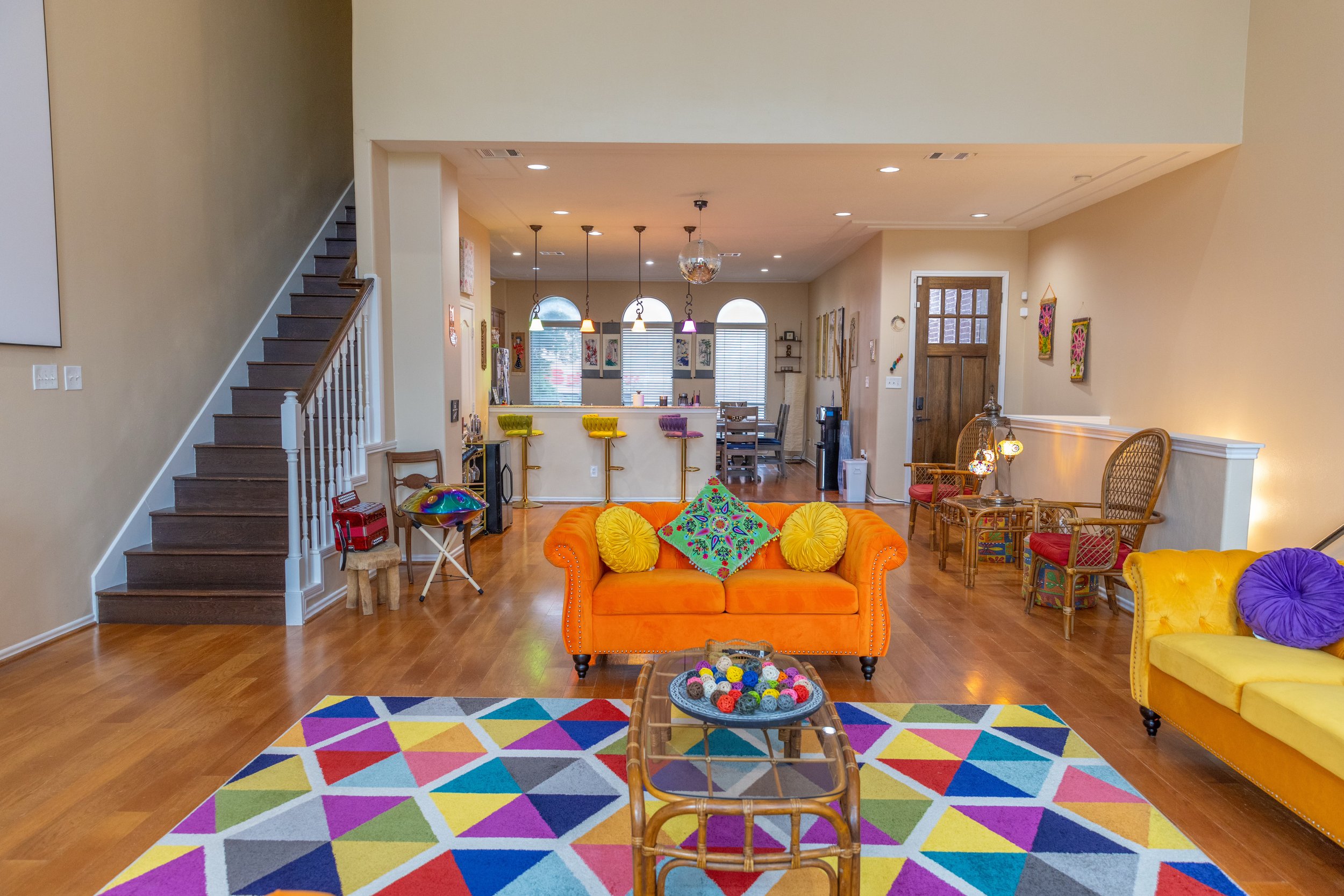 A colorful living room with orange and yellow sofas, colorful geometric rug, glass coffee table with decor, and a staircase on the left. The open room leads to a kitchen with a bar area and pendant lights. The decor is vibrant and eclectic with vario