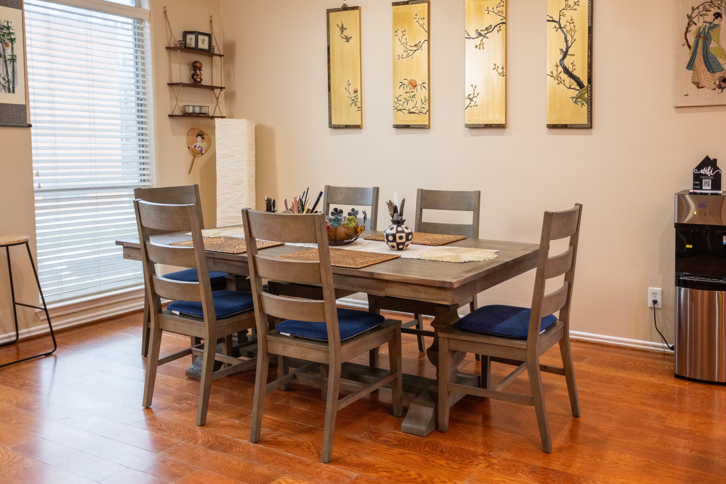 Dining room with wooden table and six chairs, art on wall, and window with blinds