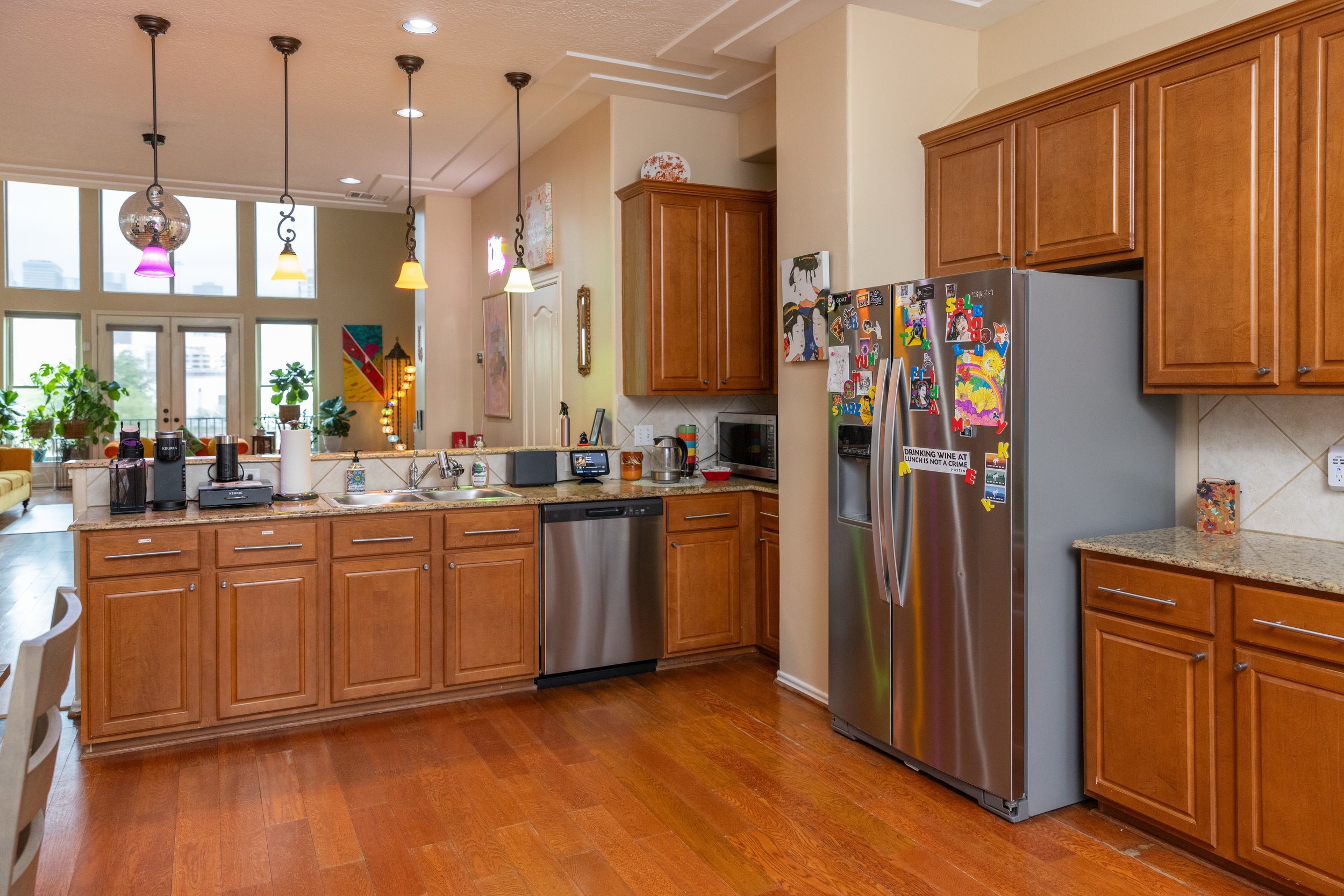 Modern kitchen interior with wooden cabinets, stainless steel appliances, colorful pendant lights, and a decorated refrigerator.