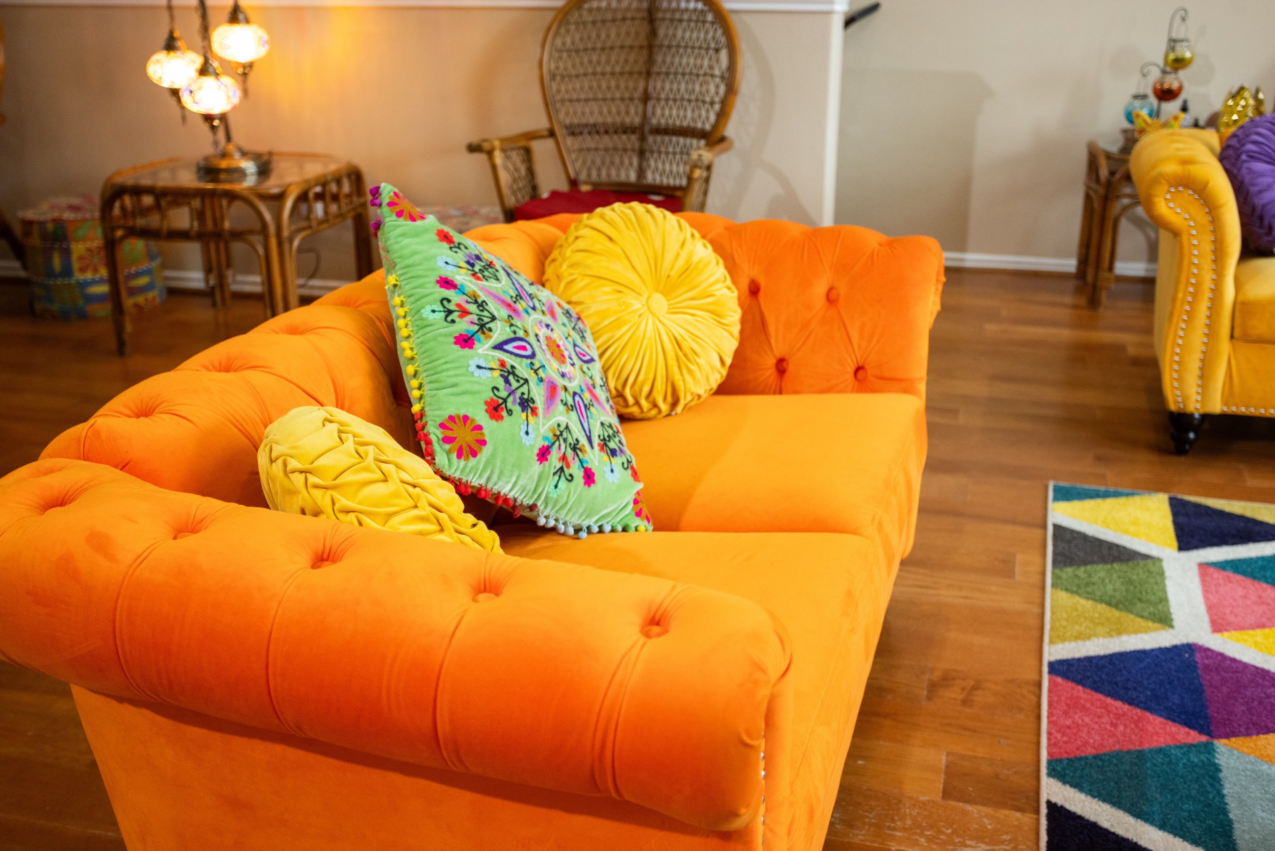 Colorful living room with an orange tufted sofa, decorative pillows, a wicker chair, a geometric patterned rug, and vintage lighting.