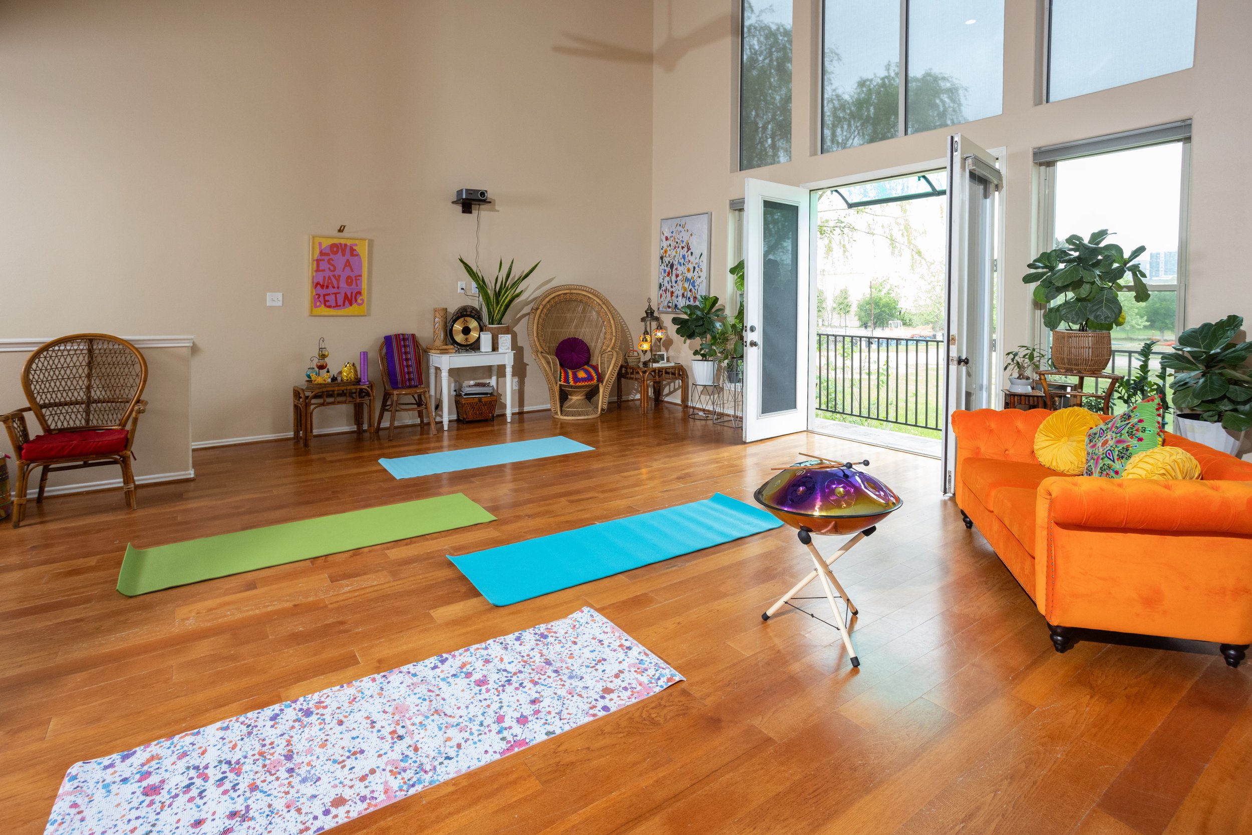 Yoga studio with colorful mats, orange sofa, musical instrument, large windows, and plants.