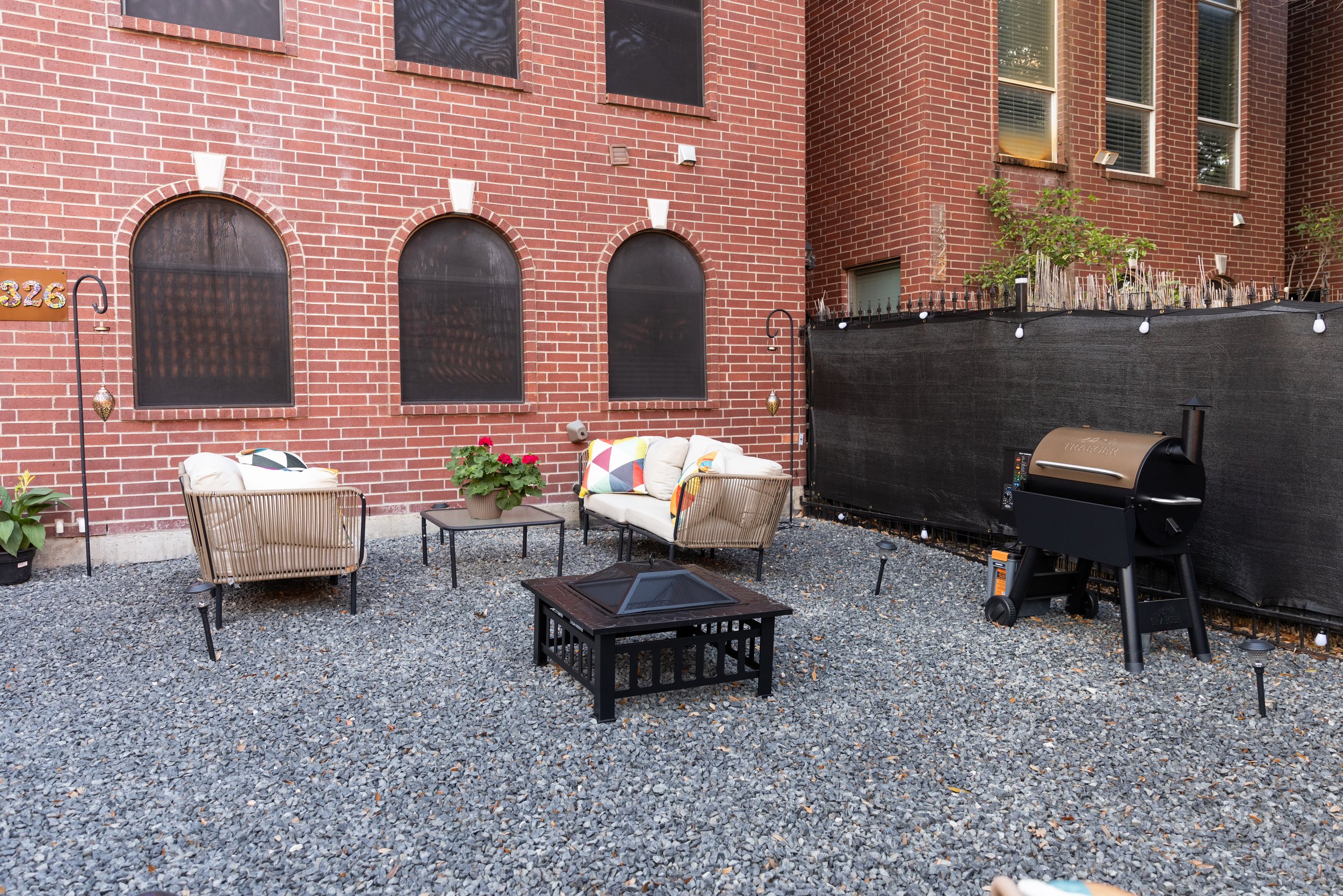 Outdoor patio with a brick wall backdrop, featuring a seating area with cushioned chairs and a table. A grill is placed next to a black privacy screen, and the ground is covered with gravel. Potted plants add greenery to the scene.