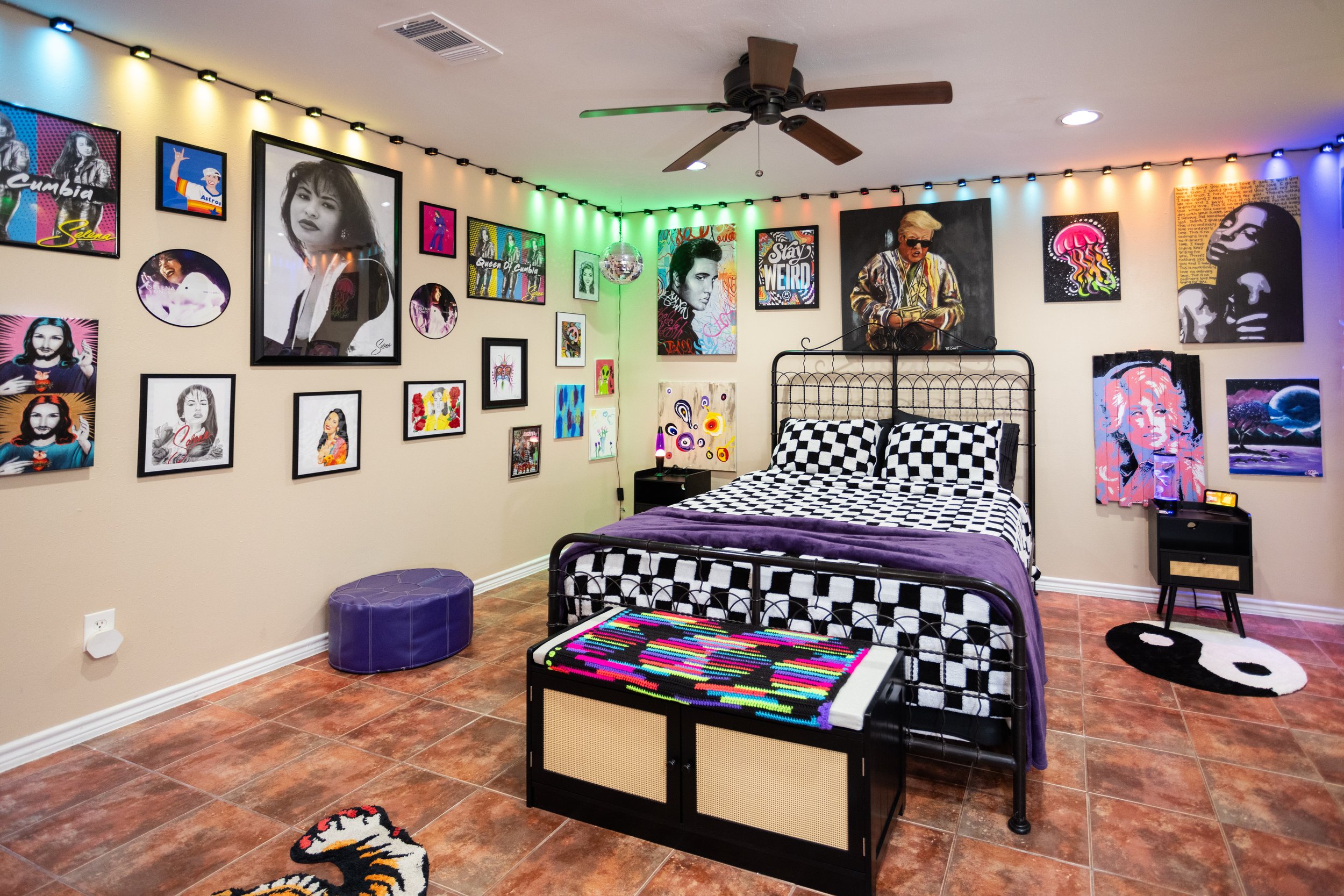A bedroom with colorful pop art and celebrity portraits on the walls, including a large black and white photo of a woman. The room features a checkered bedspread, string lights, a ceiling fan, a small bench with a colorful cushion, and a circular rug