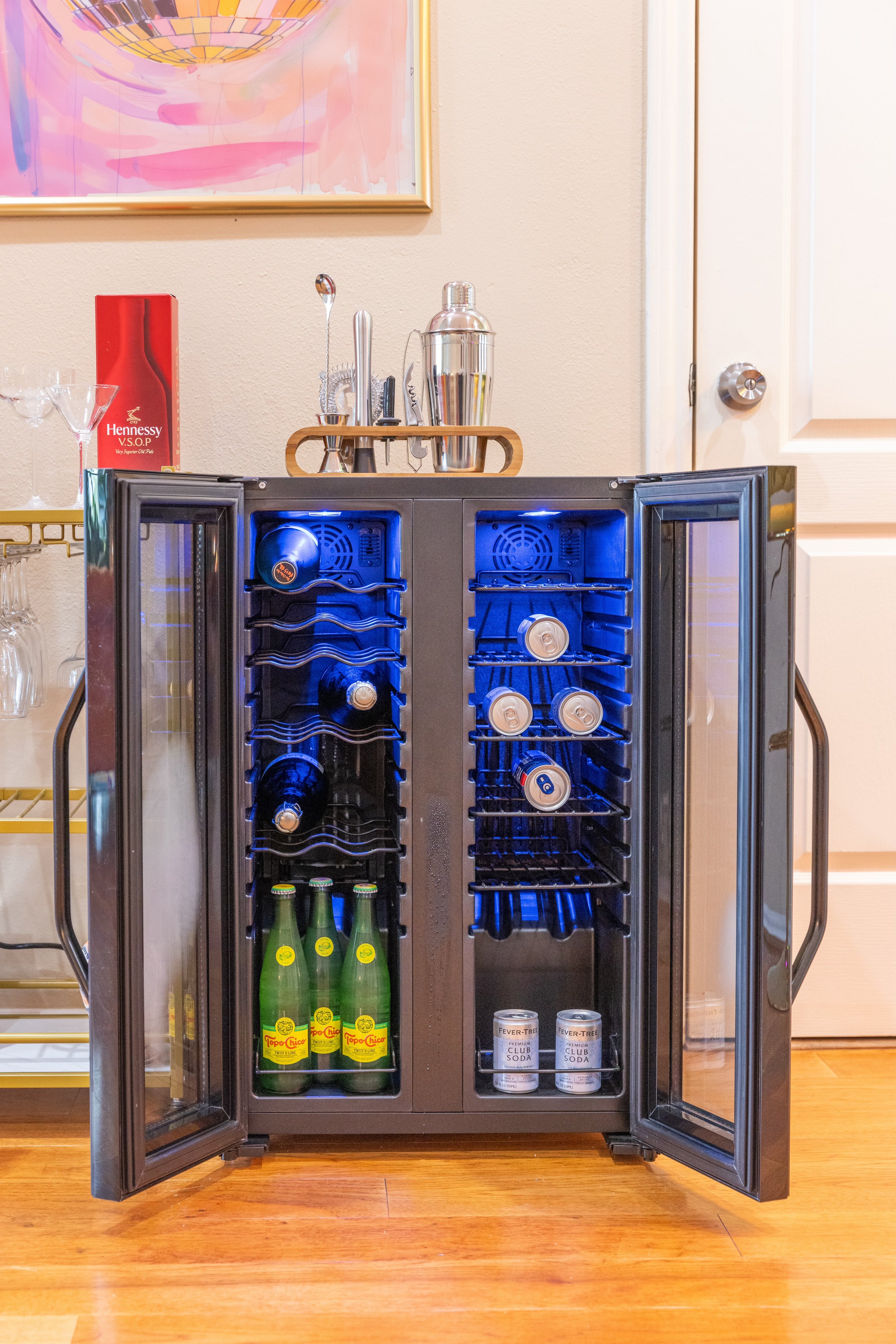 A wine and beverage cooler with open doors displaying wine bottles, cans, and bottles of sparkling water on shelves. A cocktail shaker set is on top of the cooler. The background features a bar cart and a colorful painting.