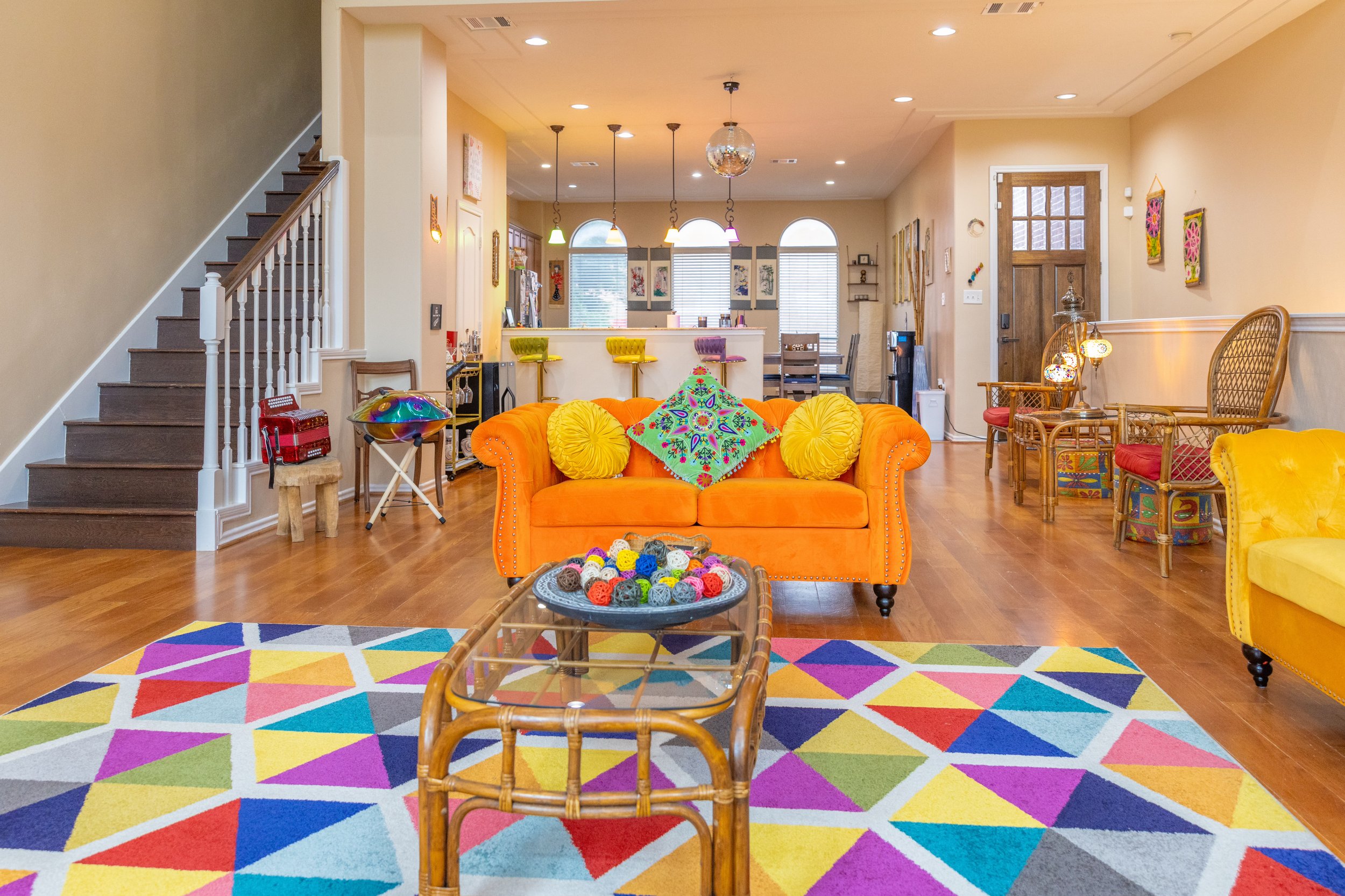 Colorful living room with orange sofa, patterned pillows, multicolored rug, and wooden flooring. Brightly decorated with various furnishings and decor items.